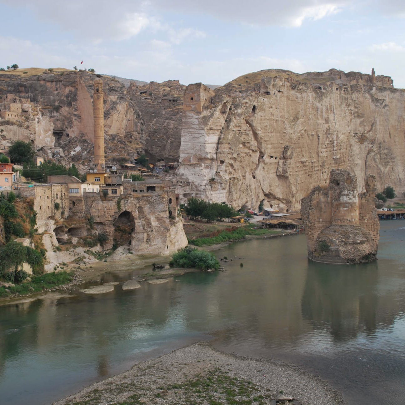 The image depicts a scenic landscape featuring a river flowing through a valley. On the left side, there are rocky cliffs and remnants of ancient structures, hinting at a historical settlement. The area appears to be a mix of natural and man-made elements, with a few buildings clinging to the hillside. In the background, there are more cliffs and a prominent tower-like structure, suggesting a rich history and possibly a fortified site. The sky is partly cloudy, adding to the serene atmosphere of the scene.