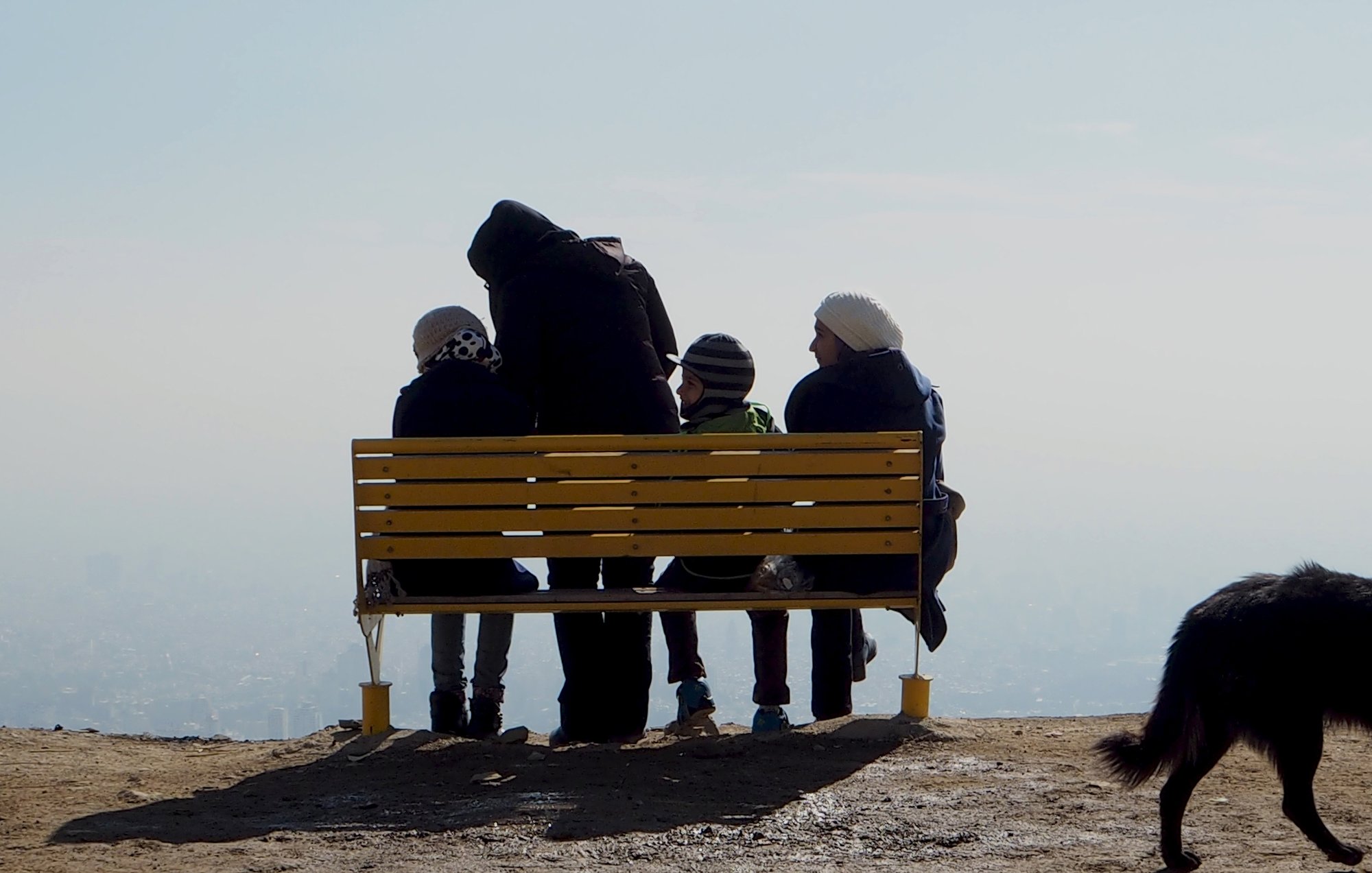 L'image montre un groupe de quatre personnes assises sur un banc jaune sur une colline ou un sommet. Ils sont tournés vers un paysage brumeux, peut-être en admirant la vue. Parmi eux, on peut voir un adulte et trois enfants. L'adulte porte un manteau sombre tandis que les enfants portent des bonnets. À droite de l'image, un chien noir se tient sur le sol, se dirigeant vers l'extérieur du cadre. L'atmosphère semble tranquille et contemplative, avec une faible visibilité en raison de la brume.