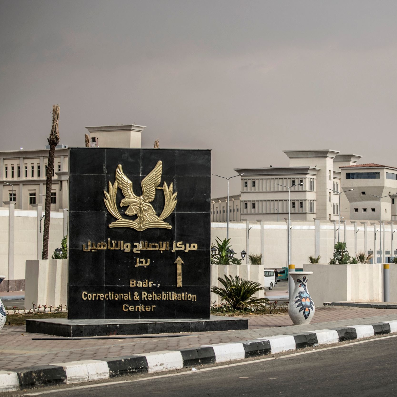 L'image montre un grand panneau d'entrée d'un centre de correction et de réhabilitation, situé à Badri, en arabe et en anglais. En arrière-plan, on peut voir des bâtiments modernes, probablement liés à ce centre. L'environnement semble urbain, avec des routes pavées et des éléments paysagers tels que des plantes. Le ciel semble légèrement nuageux, donnant une ambiance un peu sombre à la scène.