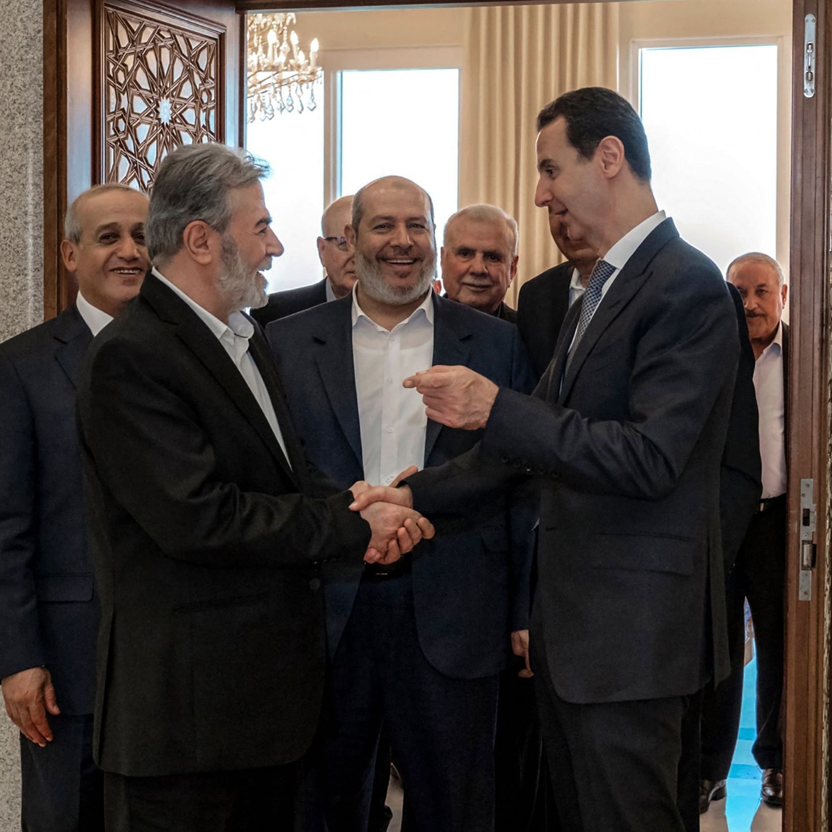 L'image montre un groupe d'hommes en train de se rencontrer dans un intérieur. Plusieurs d'entre eux sourient et semblent engagés dans une conversation amicale. L'un des hommes, au premier plan, serre la main d'un autre, tandis que les autres les observent. L'environnement semble formel, avec des éléments décoratifs visibles, comme une porte ornée et des fenêtres laissant entrer la lumière.