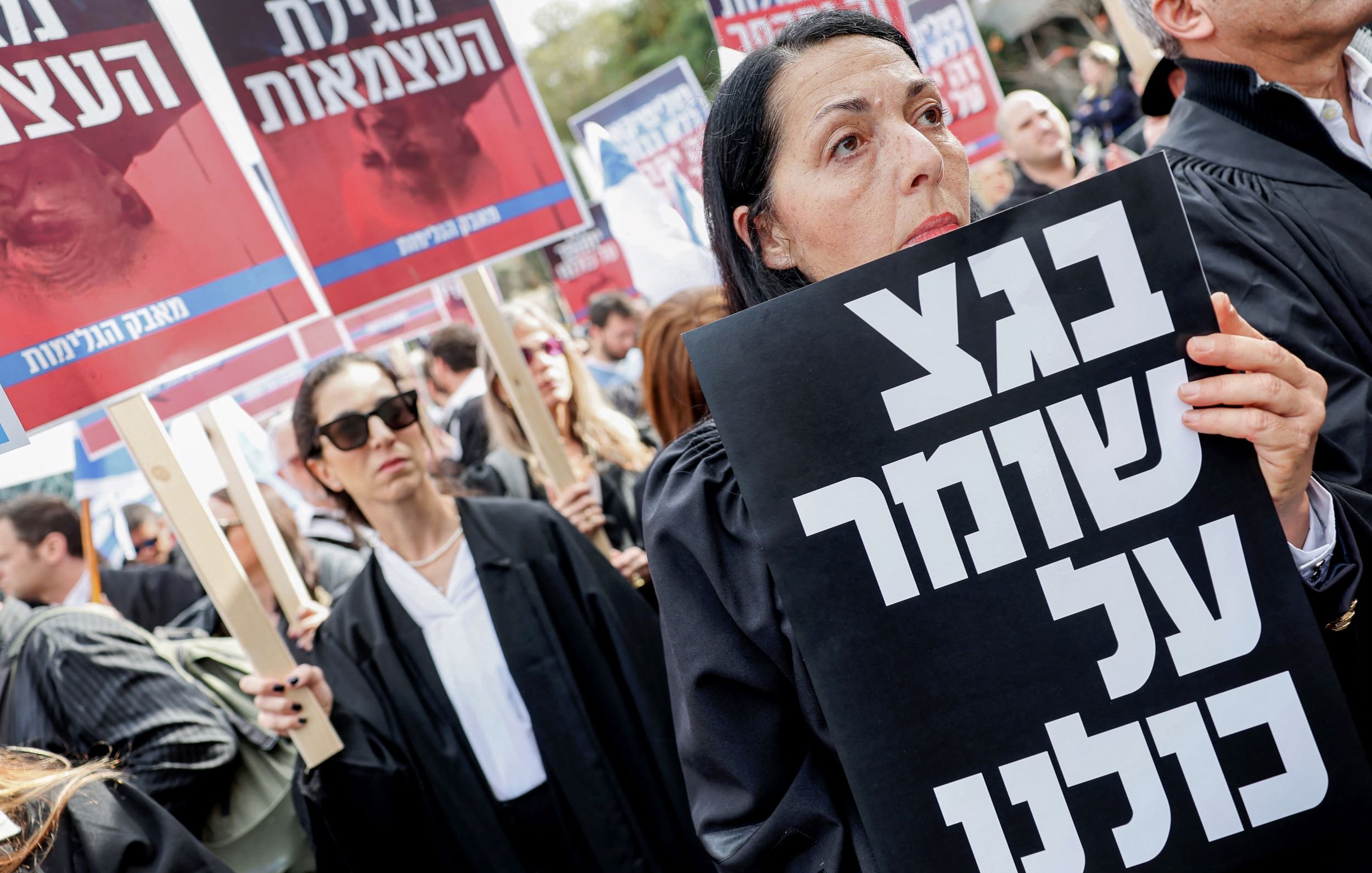 در تصویر، گروهی از افراد در حال برگزاری یک تظاهرات هستند. زنان و مردان با لباس‌های تیره و جعبه‌های دست‌نوشته‌ای در دست دارند که بر روی آنها نوشته‌هایی به زبان عبری دیده می‌شود. یکی از زنان در جلو تصویر، به وضوح در حال نگاه کردن به دوربین است و یک پلاکارد بزرگ با نوشته‌ای مشکی در دست دارد. فضای کلی تظاهرات نشان‌دهنده همبستگی و اعتراض مردم به یک موضوع خاص است.