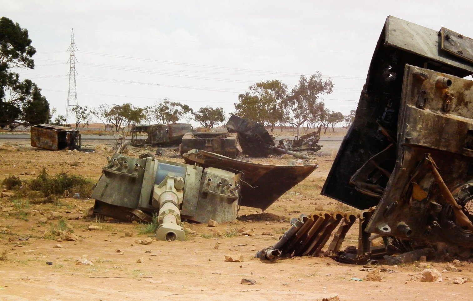 L'image montre un paysage désertique où sont dispersés des véhicules militaires rouillés et abandonnés. On peut voir des carcasses de tanks et d'autres machines, gisant sur le sol poussiéreux. Au loin, des arbres clairsemés et des pylônes électriques se distinguent, ajoutant à l'ambiance aride et désolée du lieu. Le ciel est nuageux, ce qui donne une atmosphère grise à la scène.