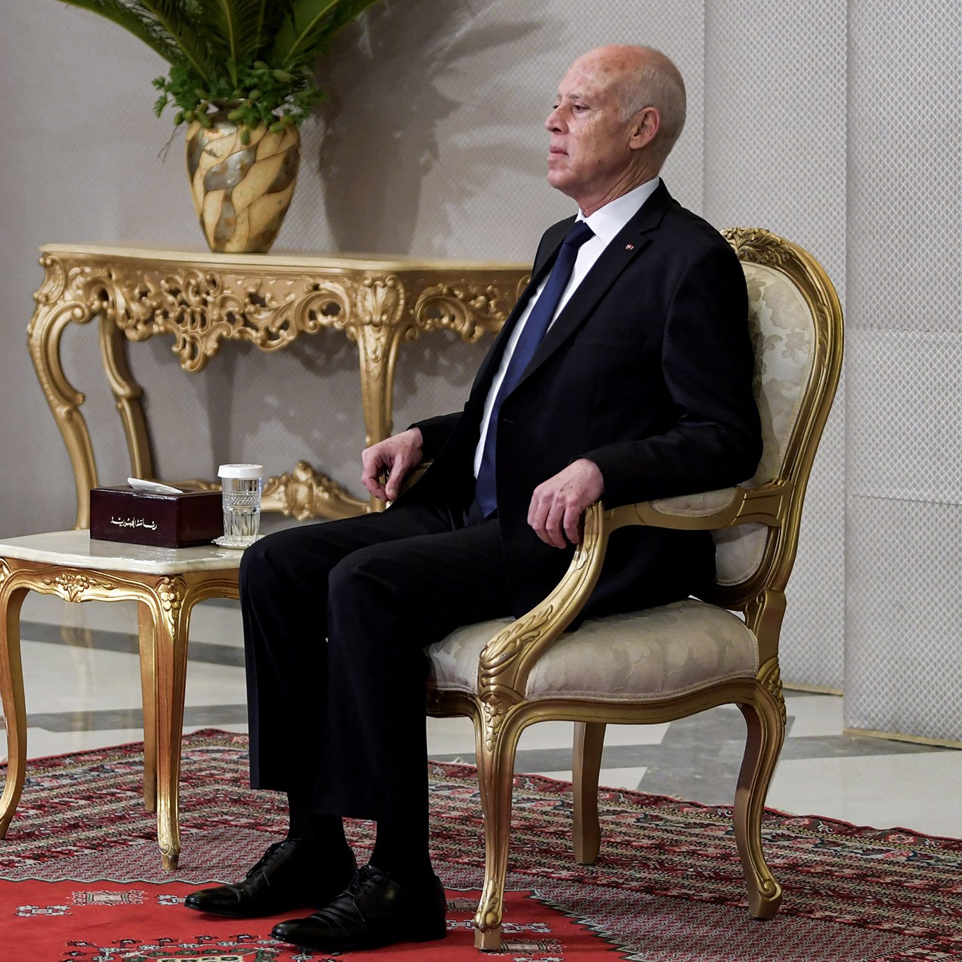 L'image montre un homme assis sur une chaise élégante dans un cadre formel. Il porte un costume noir avec une cravate. À côté de lui se trouve une table ornée sur laquelle repose une petite boîte et une tasse. L'arrière-plan présente des murs décorés et une plante en pot, ajoutant une touche de sophistication à l'environnement. Le sol est recouvert d'un tapis, contribuant à l'atmosphère formelle de la scène.