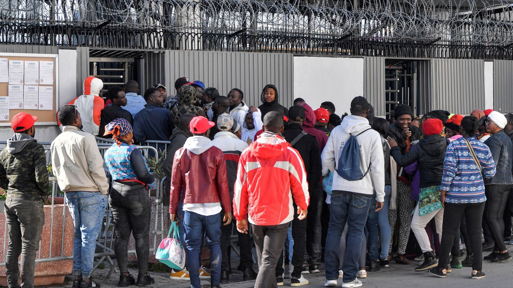 L'image montre un groupe de personnes rassemblées devant une clôture avec des barbelés. Elles semblent attendre ou essayer d'entrer dans un bâtiment. Certaines personnes portent des vêtements colorés, comme des vestes et des chapeaux rouges. Il y a une certaine densité dans la foule, suggérant une situation de rassemblement ou de file d'attente. Le mur en arrière-plan comporte des affichages et il y a une ambiance de tension ou d'importance autour de l'endroit où se trouvent les personnes.