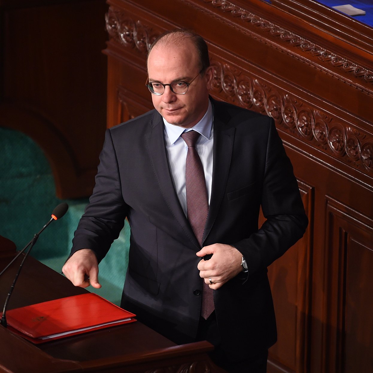 L'image montre un homme en costume sombre, portant des lunettes, se tenant près d'un pupitre dans une assemblée ou un parlement. Il semble être en train de s'adresser à l'assemblée ou de faire une déclaration informative, avec un dossier rouge posé sur le pupitre. Le décor est constitué de boiseries et de sièges verts, typiques d'un environnement institutionnel.