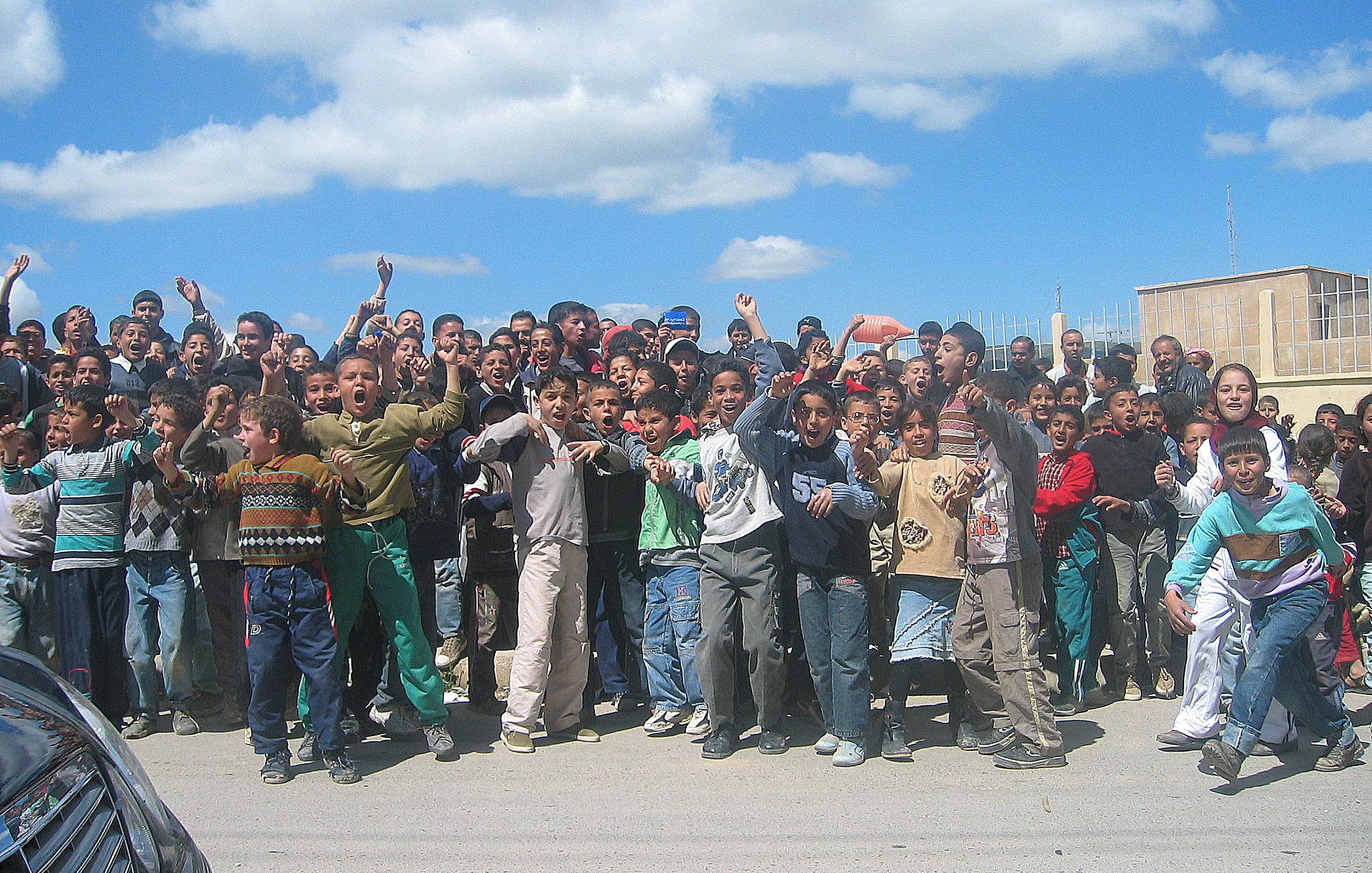 L'image montre un groupe important d'enfants et d'adolescents rassemblés en plein air. Ils semblent joyeux et excités, levant les bras et faisant des gestes. Le ciel est clair avec quelques nuages, et l'environnement urbain en arrière-plan suggère un cadre scolaire ou communautaire. Les enfants portent des vêtements variés, reflet de leur diversité. L'atmosphère semble animée et festive.