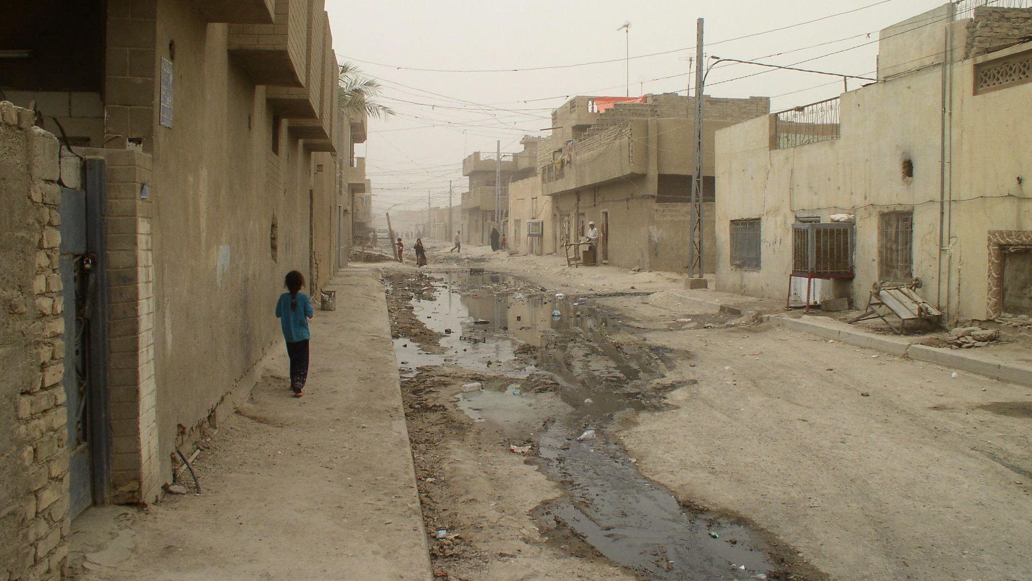 الصورة تظهر شارعًا في منطقة سكنية تبدو مهملة، حيث تظهر المباني ذات اللون الرمادي والخالي من الزينة. الطريق يبدو غير معبد ويظهر فيه أجزاء من المياه والأتربة. هناك شخصان يسيران في الشارع، ورغم الغبار، فإن تفاصيل الحياة اليومية تظهر في هذه البيئة. الشارع يبدو ضيقًا وفارغًا، مما يعكس حالة من الإهمال والبساطة في التصميم العمراني.