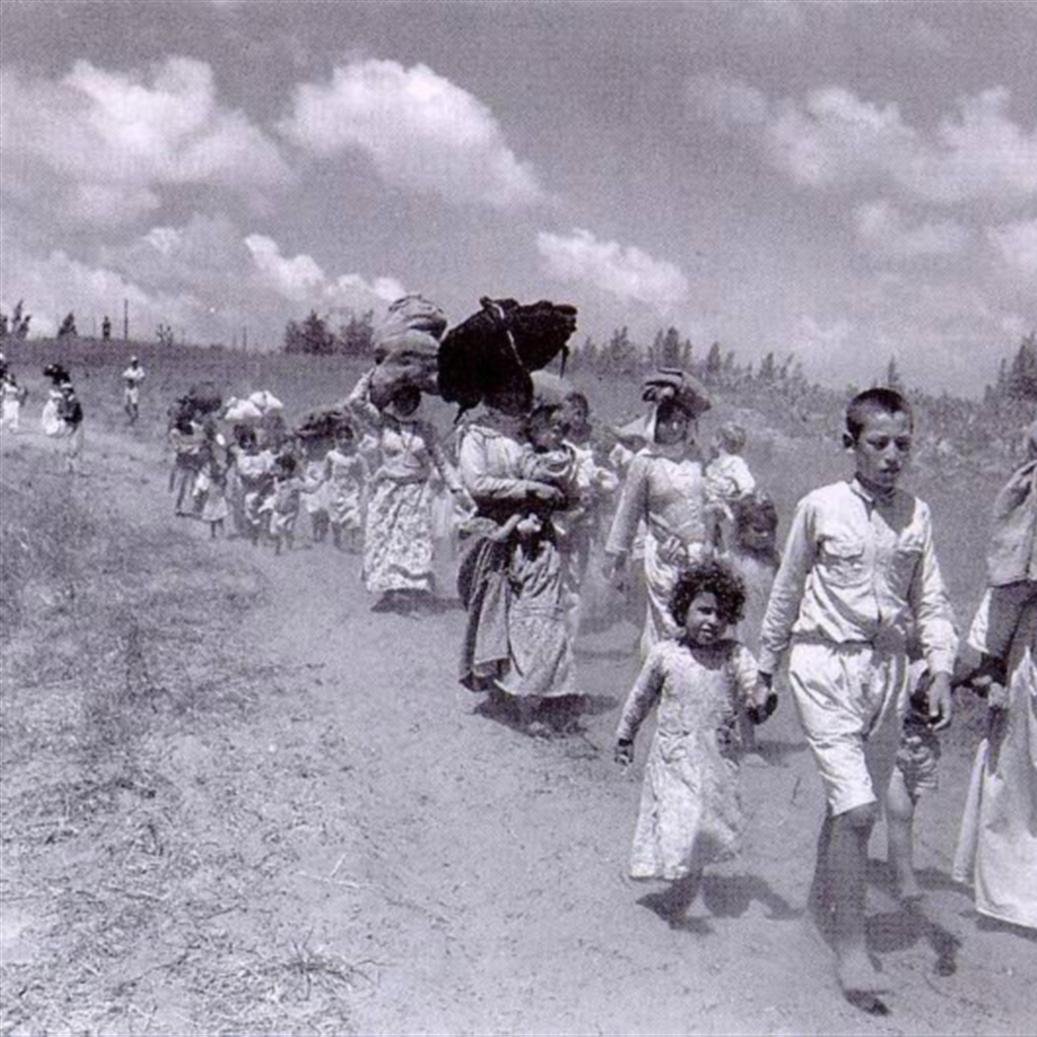 L'image montre un groupe de personnes marchant sur un chemin. On y voit des hommes, des femmes et des enfants, portant des charges sur leur tête ou dans leurs bras. Les adultes semblent laborieux, tandis que les enfants marchent à leurs côtés. Le paysage est rural, avec un ciel nuageux en arrière-plan, et l'atmosphère générale évoque une situation de déplacement ou d'exode. Les vêtements des personnes sont simples et évoquent une époque passée.