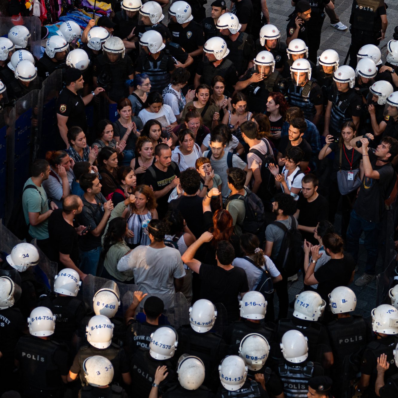 L'image montre une scène de manifestation où une foule de personnes se trouve face à des policiers en tenue anti-émeute. Les manifestants semblent discuter ou protester, tandis que les policiers forment une ligne serrée. L'atmosphère est tendue, et une partie de la foule semble concentrée sur un point, indiquant une interaction ou un affrontement verbal. Les casques des policiers et leur équipement renforcent l'impression de confrontation.