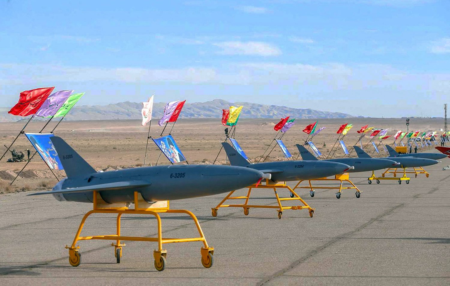 L'image montre une série de drones militaires disposés sur une piste. Les drones sont de couleur grise et sont montés sur des chariots jaunes. Au-dessus de chaque drone, il y a des drapeaux colorés qui flottent dans le vent. En arrière-plan, le paysage est désertique, avec des collines et des montagnes au loin, sous un ciel clair. Cette scène semble être une présentation ou un défilé militaire.