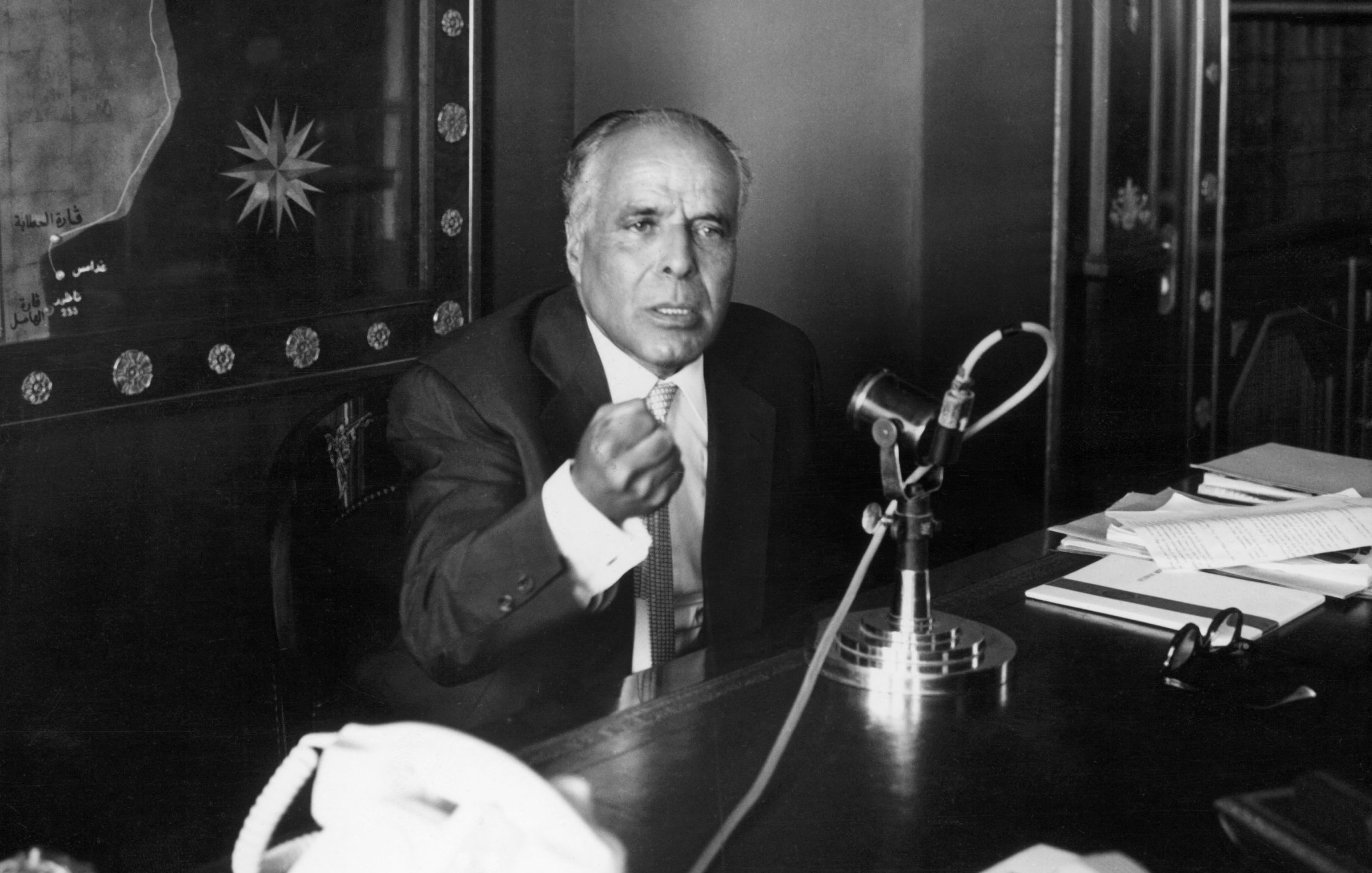 Homme en costume, gesticulant, devant un microphone et un bureau rempli de documents.