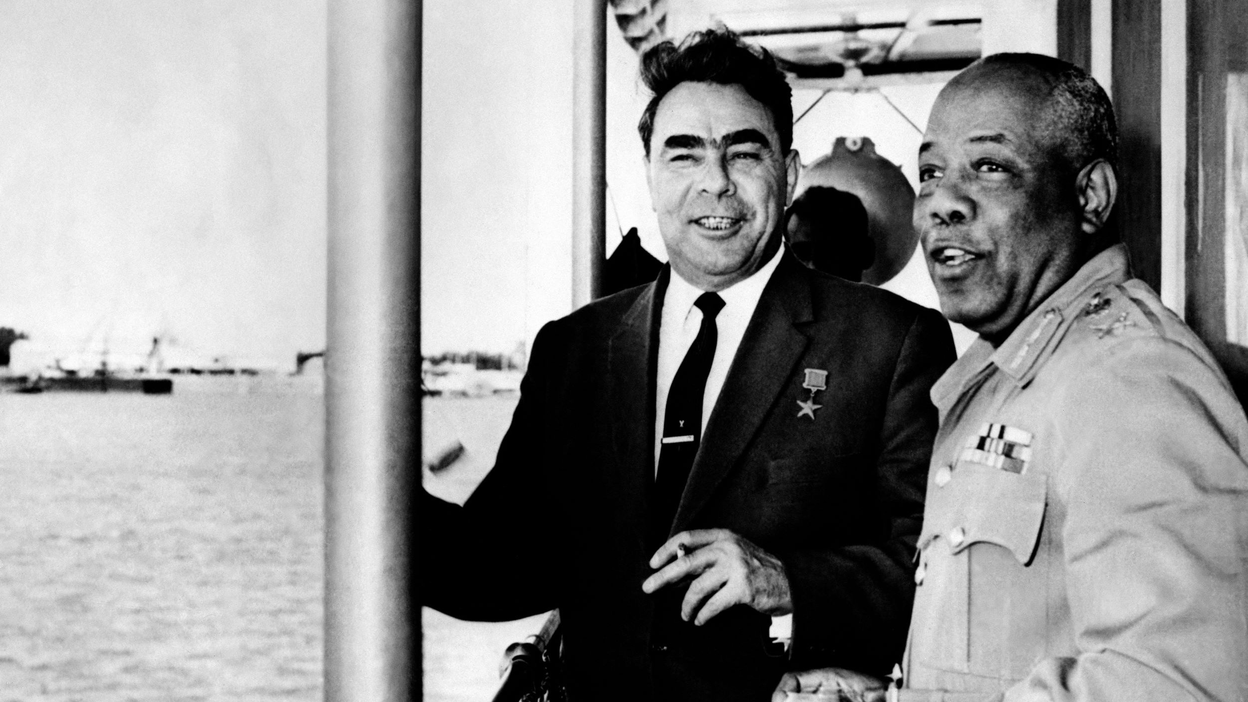 L'image montre deux hommes en train de discuter sur ce qui semble être un bateau ou une plateforme près de l'eau. L'un des hommes porte une médaille, indiquant honneur ou reconnaissance, et les deux semblent souriants et détendus. En arrière-plan, on peut voir des bateaux et une vue maritime. L'atmosphère générale de l'image semble amicale et conviviale.