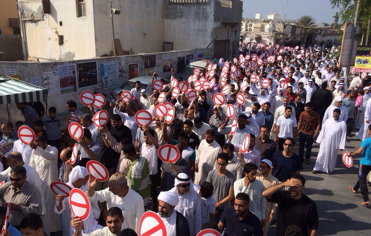 الصورة تظهر مجموعة كبيرة من الناس في مظاهرة أو احتجاج. المشاركون يحملون لافتات كبيرة عليها إشارات تعبر عن ممنوع أو خط أحمر. يرتدي الكثير منهم ملابس تقليدية، وتبدو الحشود منظمة. الخلفية تحتوي على مبانٍ وصور ملصقة على الجدران، مما يشير إلى أن الحدث يحدث في منطقة حضرية.