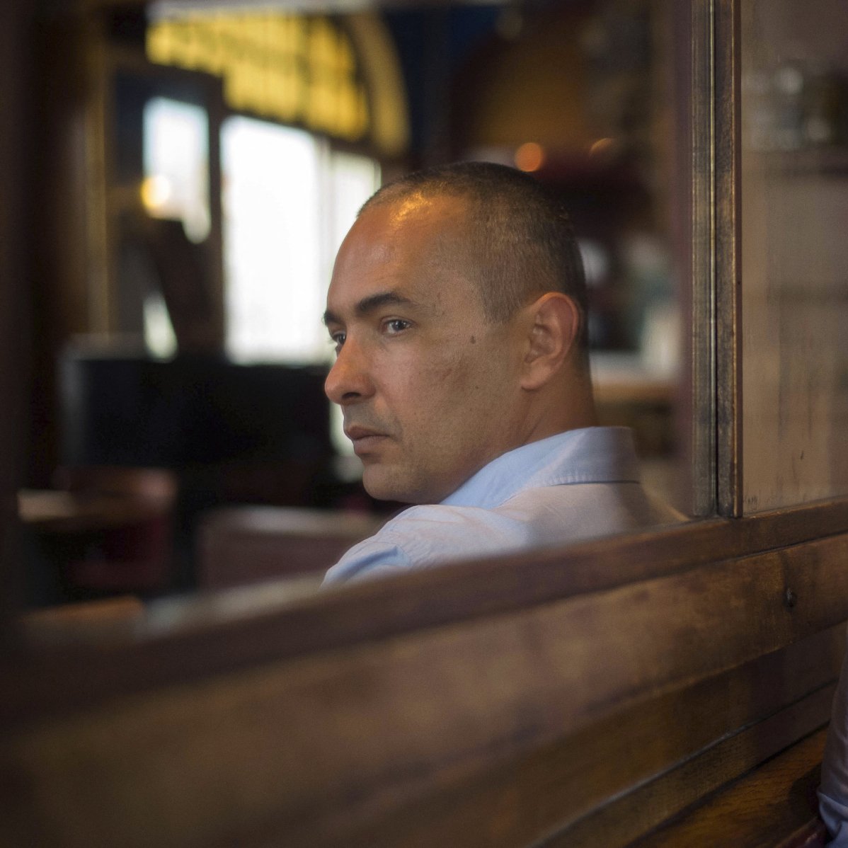 تظهر الصورة شخصًا يجلس على مقعد خشبي، مع انعكاسه في زجاج خلفه. يبدو أن الرجل في منتصف العمر، لديه شعر قصير ومظهر هادئ. يرتدي قميصًا أزرق فاتح، وينظر مباشرة إلى الكاميرا بتعبير جاد. الخلفية غير واضحة، مما يركز الانتباه على الشخص ووجهه. الإضاءة في الصورة دافئة وتساهم في خلق جو من التأمل والعمق.