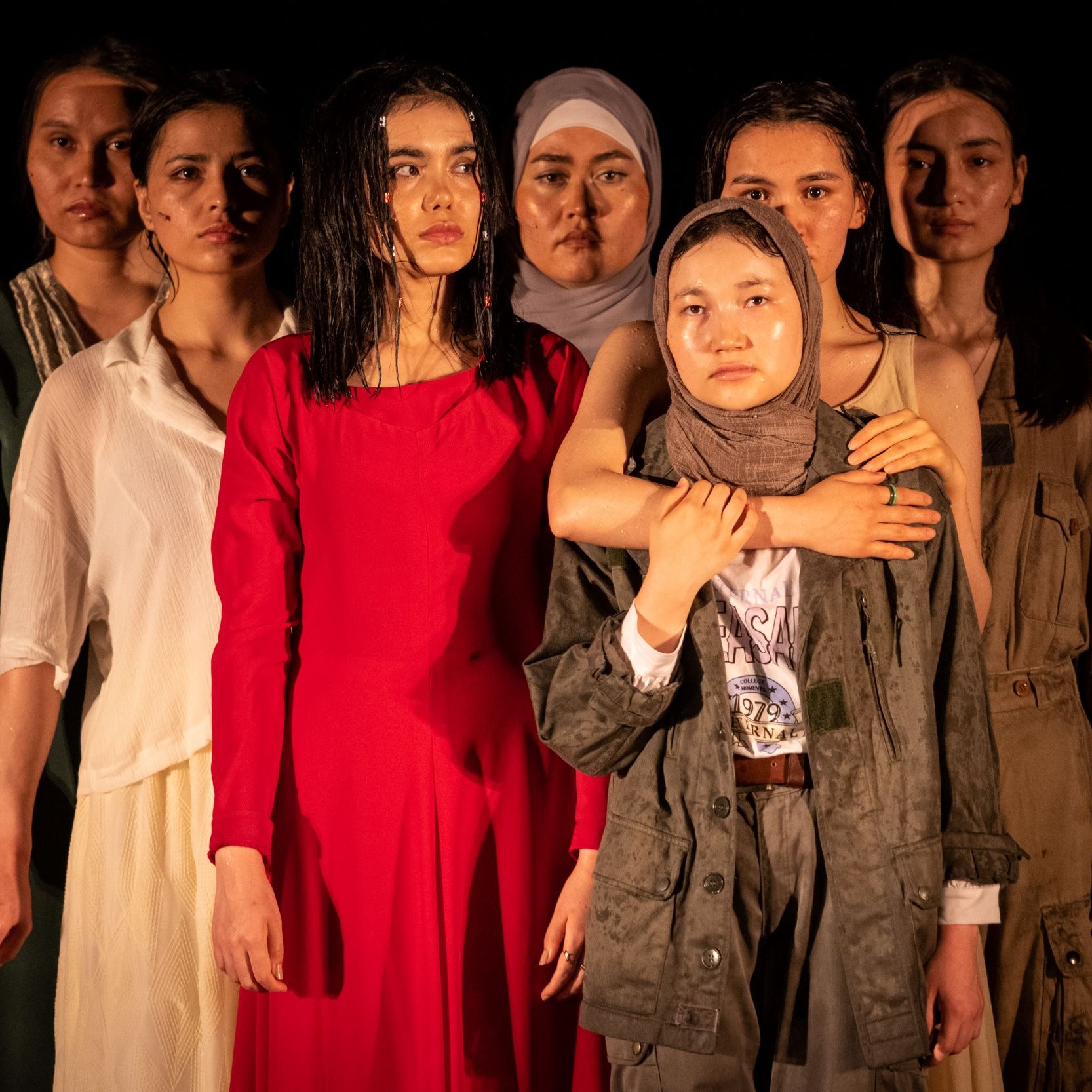 L'image montre un groupe de sept femmes, chacune portant des vêtements différents. Elles se tiennent en rang, avec des expressions sérieuses sur leurs visages. La femme au centre, vêtue d'une robe rouge, attire particulièrement l'attention. Les autres femmes, portant des vêtements variés allant de styles modernes à plus traditionnels, affichent une diversité de couleurs et de textures. L'éclairage met en valeur leurs visages et leurs postures, créant une atmosphère à la fois forte et émotive.