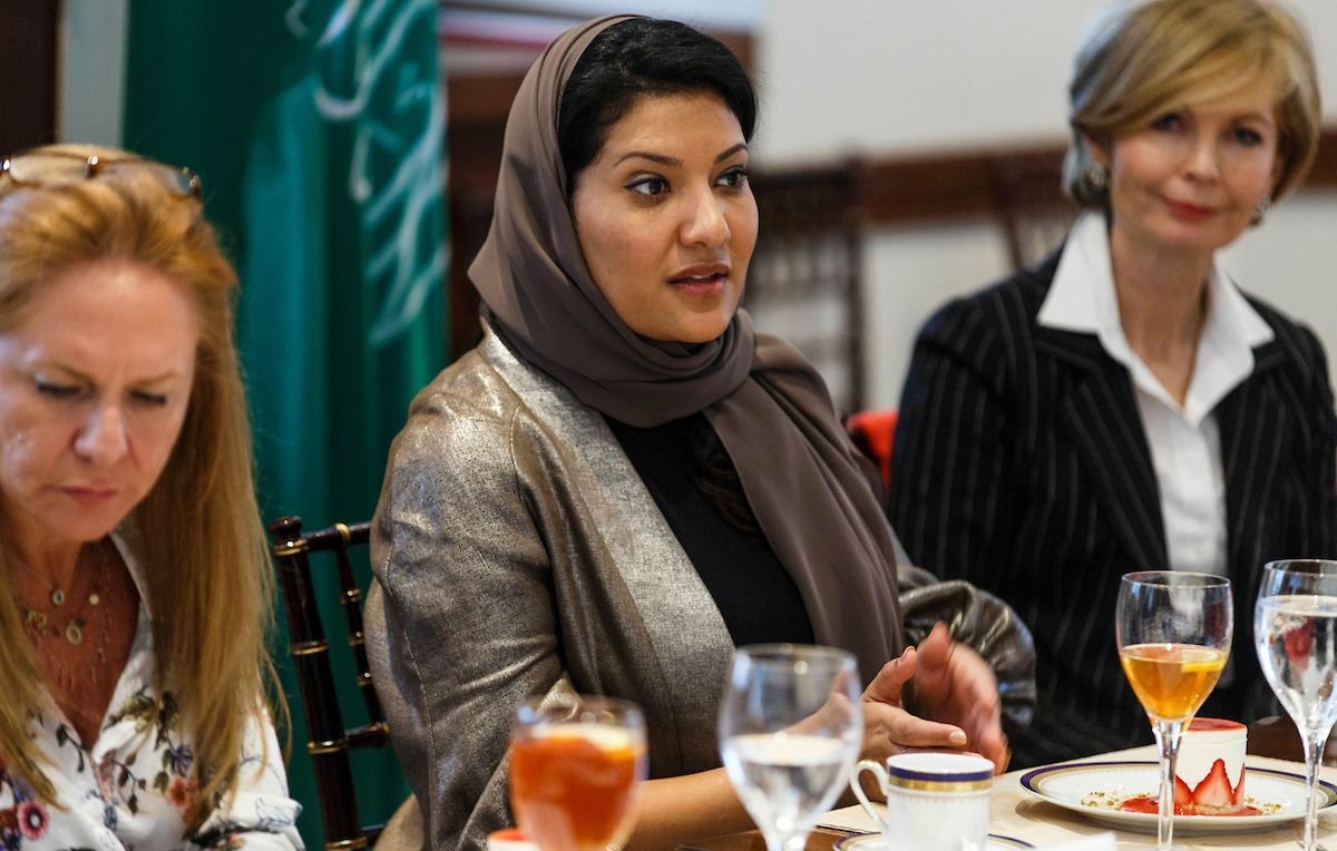 L'image montre un groupe de femmes assises autour d'une table, en train de discuter. Une femme au centre porte un hijab et un vêtement aux couleurs neutres, semblant engagée dans la conversation. À sa gauche, une autre femme aux cheveux longs et blonds, légèrement penchée, écoute attentivement. À droite, une femme en tailleur noir et blanc observe également. Sur la table, on peut voir des verres de boisson et de la vaisselle, avec des éléments de nourriture. L'environnement semble formel, propice à une réunion ou un déjeuner d'affaires.