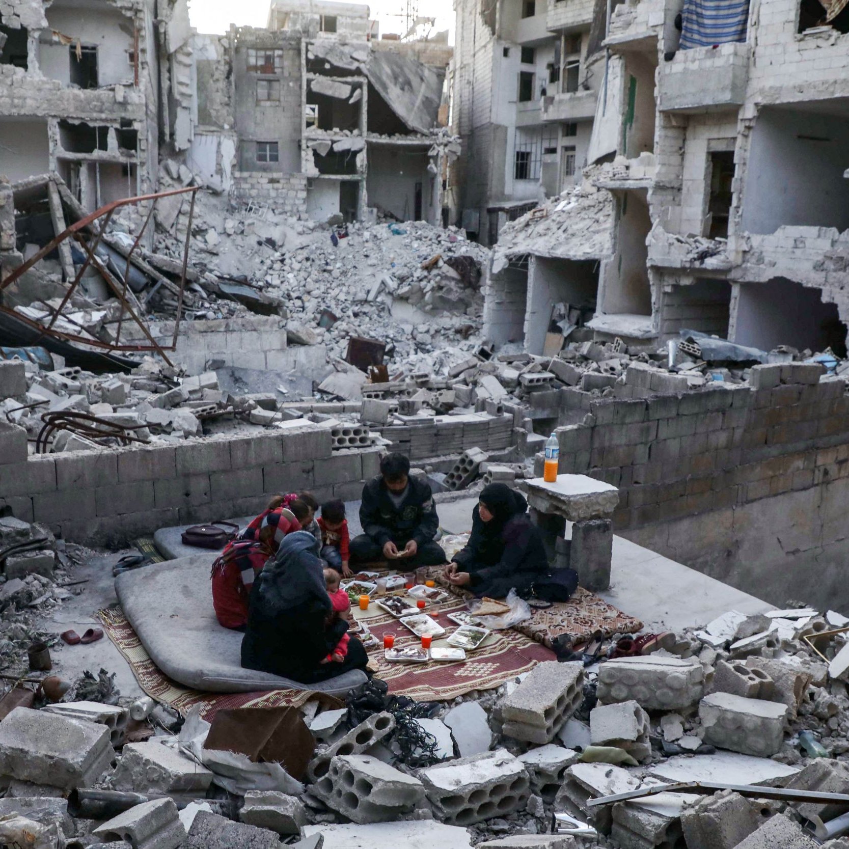 L'image montre un groupe de personnes assises sur un tapis dans un environnement dévasté. Autour d'eux, on peut voir des débris de bâtiments effondrés et des structures endommagées. Les personnes semblent concentrées sur une activité, malgré la destruction qui les entoure. L'atmosphère évoque la résilience et la vie qui continue malgré les difficultés.