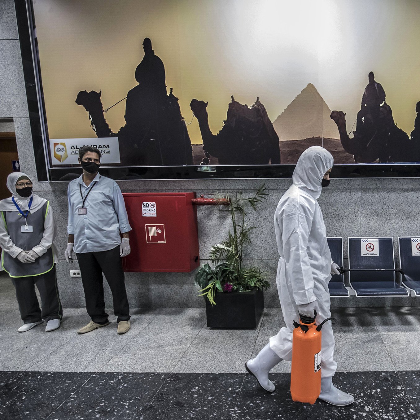 در این تصویر، یک فرد در حال قدم زدن است که لباسProtective پوشیده و یک جعبه نارنجی رنگ در دست دارد. در پس‌زمینه، تصویری از اهرام مصر و افرادی که بر روی شترها نشسته‌اند، به صورت سیاه و سفید دیده می‌شود. همچنین، دو نفر دیگر در سمت چپ تصویر ایستاده‌اند و ماسک زده‌اند. فضای کلی تصویر گویا محیطی عمومی یا فرودگاهی است.