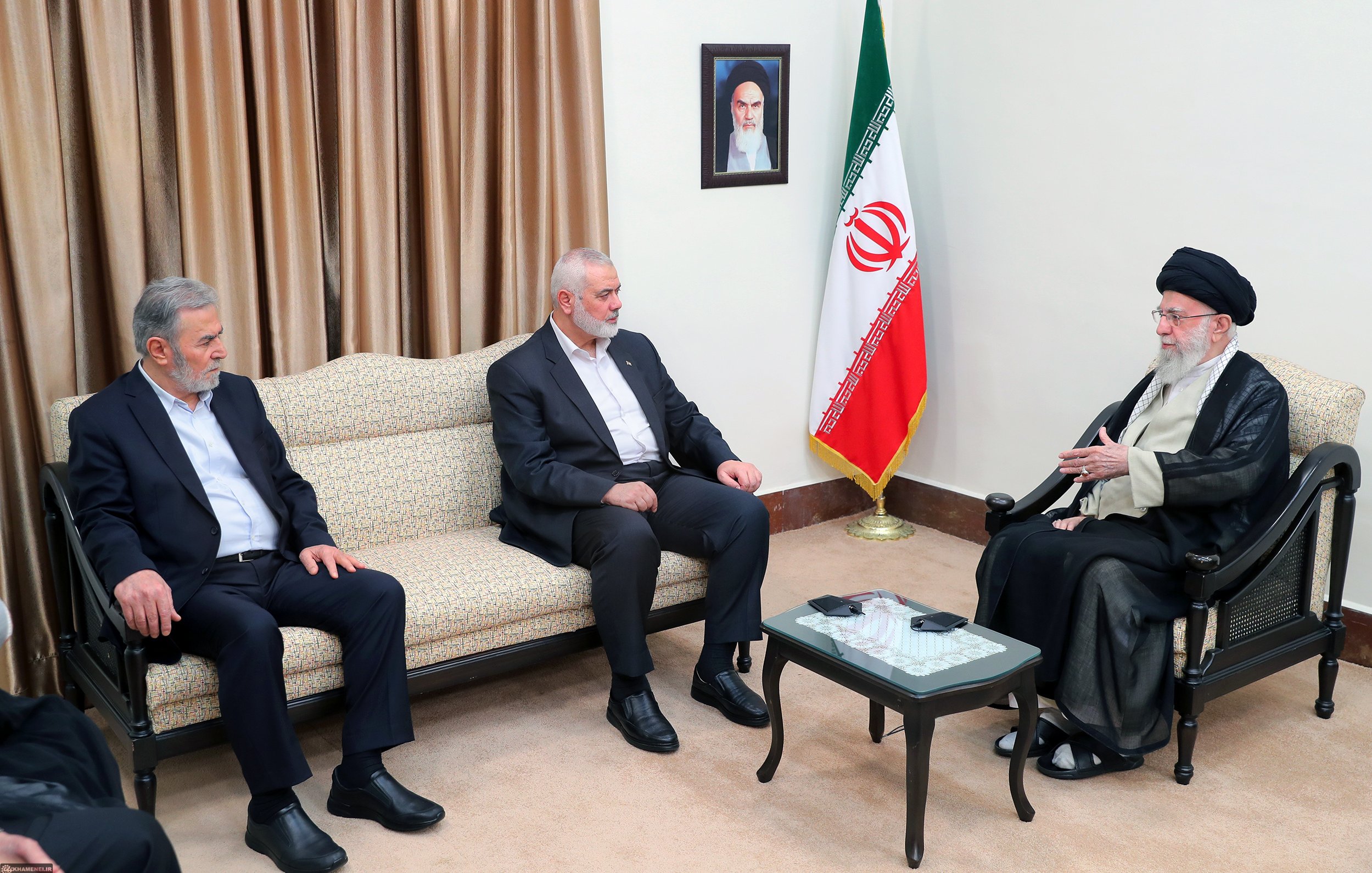 L'immagine mostra un incontro tra tre uomini. Due di loro sono seduti su un divano, mentre il terzo, che indossa un abbigliamento tradizionale, è seduto di fronte a loro. Sul muro si trova un ritratto e, accanto, è presente una bandiera iraniana. L'atmosfera sembra formale e il gruppo appare impegnato in una discussione.