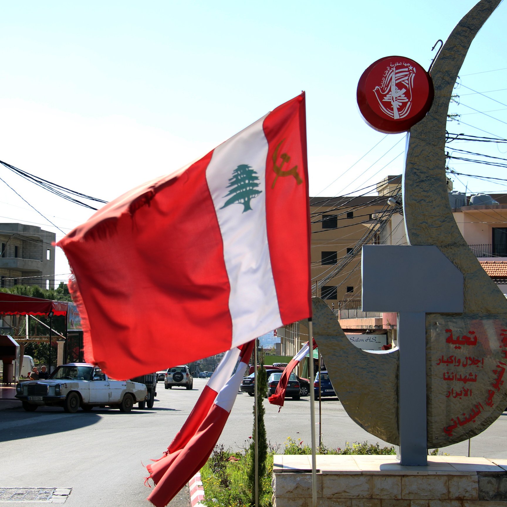 L'image représente un paysage urbain ensoleillé au Liban. On y voit un drapeau libanais flottant au vent, avec une grande emblème en métal en arrière-plan. Cet emblème est orné de symboles et de signification culturelle, illustrant probablement l'identité locale ou nationale. Les bâtiments autour semblent typiques de la région, avec des lignes électriques visibles et un ciel clair en arrière-plan.