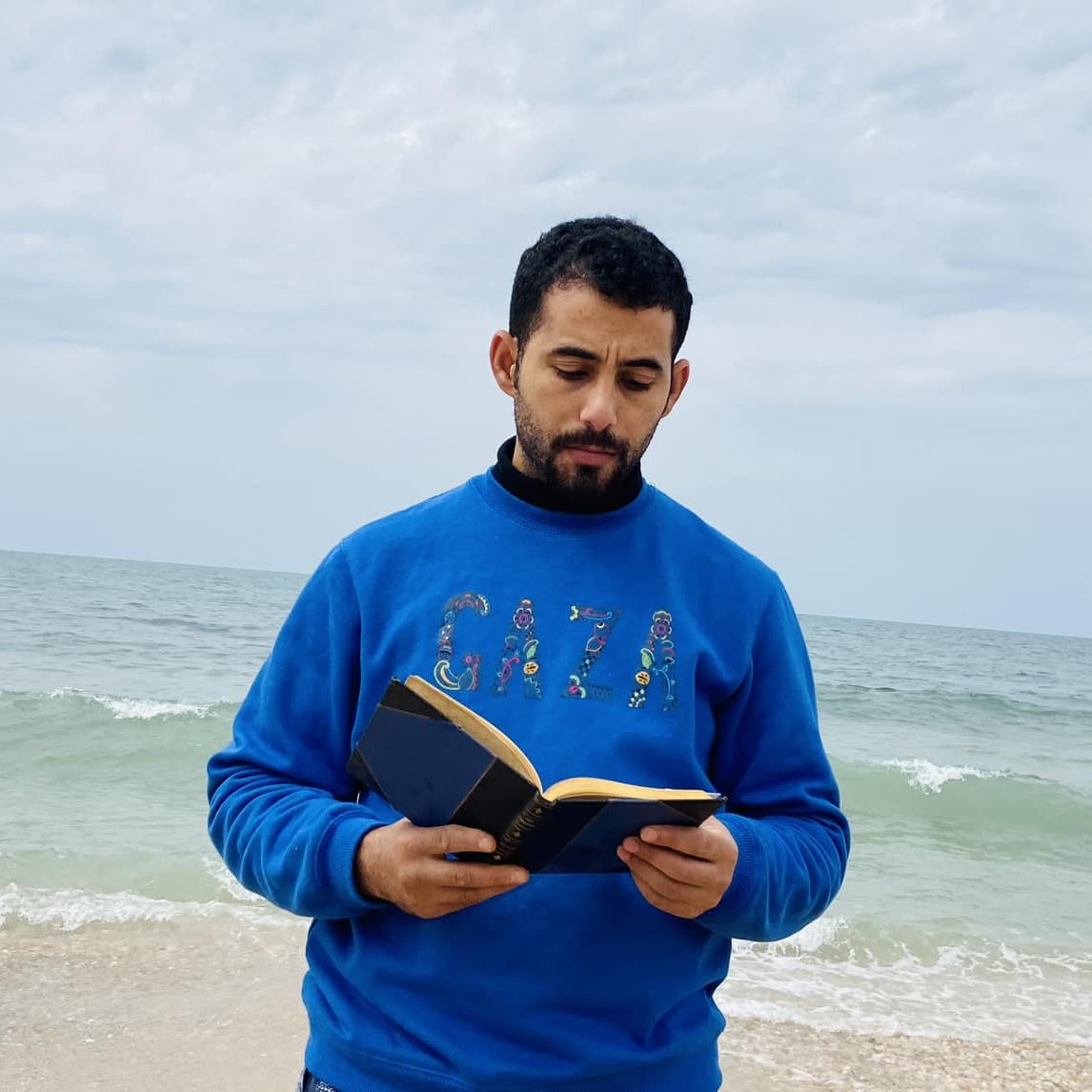 L'image montre un homme debout sur une plage, face à la mer. Il porte un pull bleu avec "GAZA" écrit dessus. Il tient un livre ouvert dans ses mains et semble lire ou se concentrer sur son texte. En arrière-plan, on peut voir des vagues qui se brisent doucement sur le rivage, et le ciel est nuageux, ajoutant une atmosphère calme à la scène.