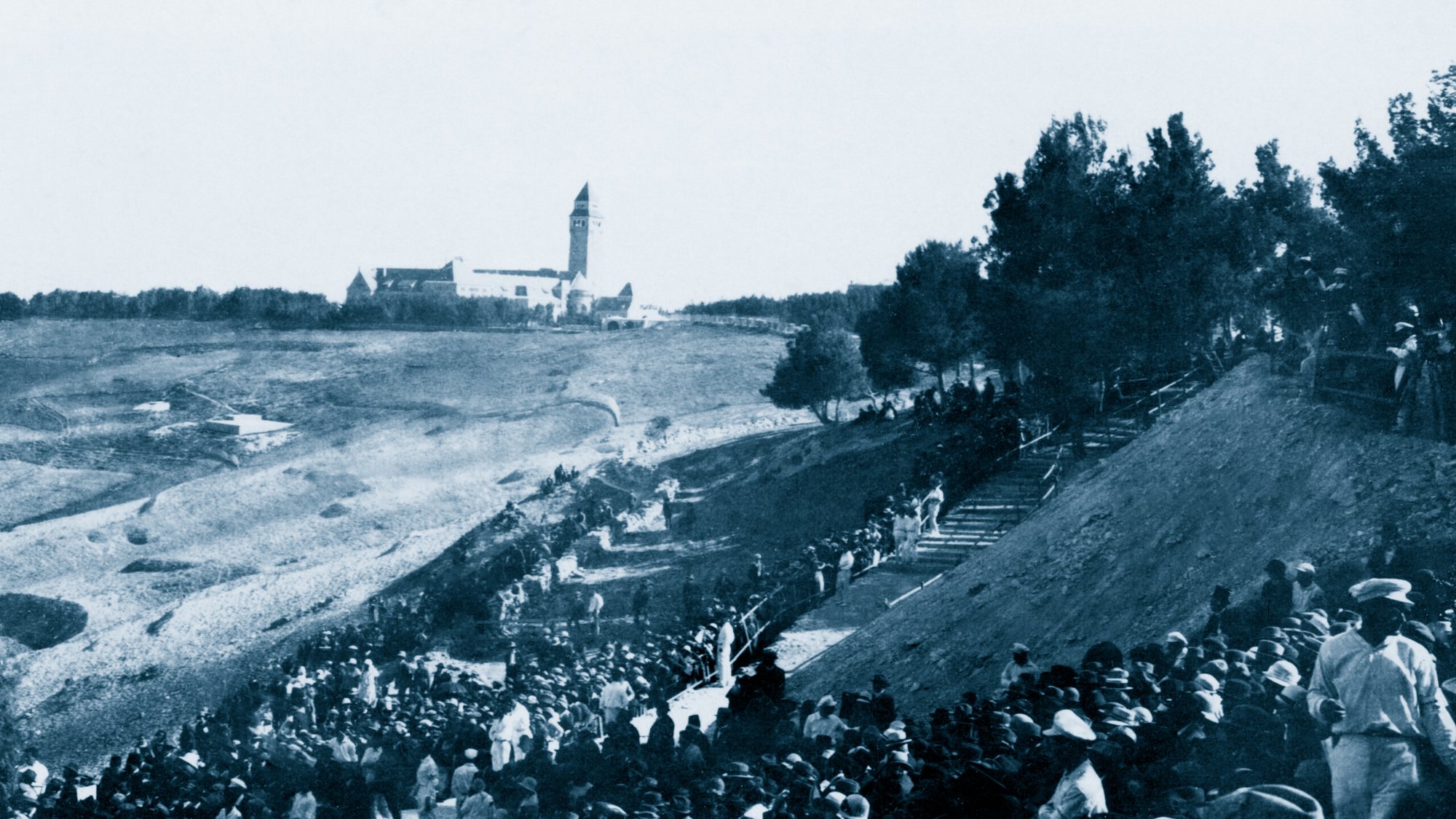A large crowd in historical clothing gathers on a hillside, with a building in the distance.