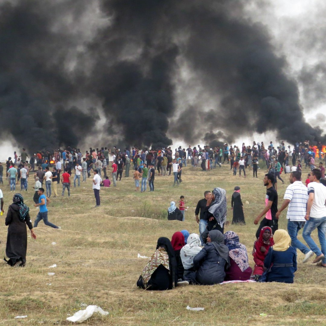L'image montre une grande foule de personnes rassemblées sur une colline, avec d'importants nuages de fumée noire émanant en arrière-plan. Les gens semblent se tenir debout ou marcher, certains regardant vers la fumée. Au premier plan, un groupe de personnes est assis sur le sol, tandis que d'autres se déplacent autour d'eux. L'ambiance est tendue, indiquant une situation d'agitation ou de protestation. Le paysage est principalement sec avec quelques herbes.