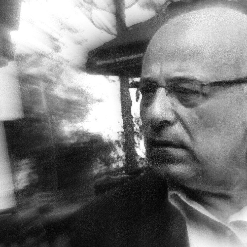 تظهر الصورة رجلًا في منتصف العمر، يبدو جادًا ومتأملًا. يعبر وجهه عن التفكير العميق أو الانشغال بشيء ما. يبدو أن الصورة قد أُخذت في مكان خارجي، حيث يمكن رؤية بعض العناصر الطبيعية أو المعمارية في الخلفية، مما يعطي إحساسًا بالعمق. الصورة تبدو مشوشة قليلاً، مما يضيف تأثيرًا فنيًا إليها.