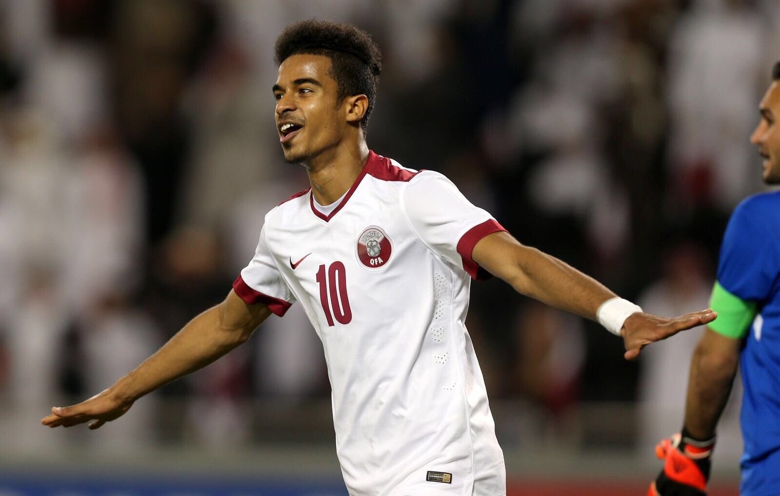 L'image montre un jeune joueur de football qui célèbre un but. Il porte un maillot blanc avec des touches de rouge, et le numéro 10 est visible sur son dos. Son expression est joyeuse et dynamique, manifestant sa satisfaction après une action réussie. En arrière-plan, on aperçoit des supporters, probablement vêtus de blanc, ce qui suggère un événement sportif important. La scène dégage une énergie positive et une intensité propre au football.