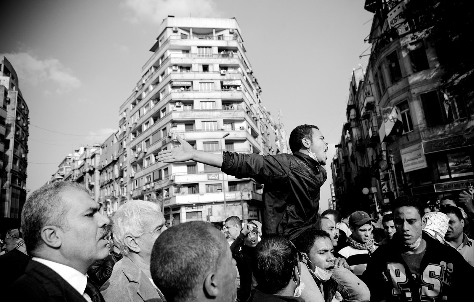 L'image montre une scène de manifestation dans un environnement urbain. Au centre, un homme se tient debout, les bras écartés, semblant exprimer une idée forte ou appeler à l'action. Il est entouré d'une foule dense, composée d'hommes, qui l'écoute ou réagit à son discours. Les bâtiments en arrière-plan montrent une architecture typique de ville, et l'image est en noir et blanc, ce qui accentue les émotions et la tension de la situation. L'atmosphère semble dynamique et engageante.
