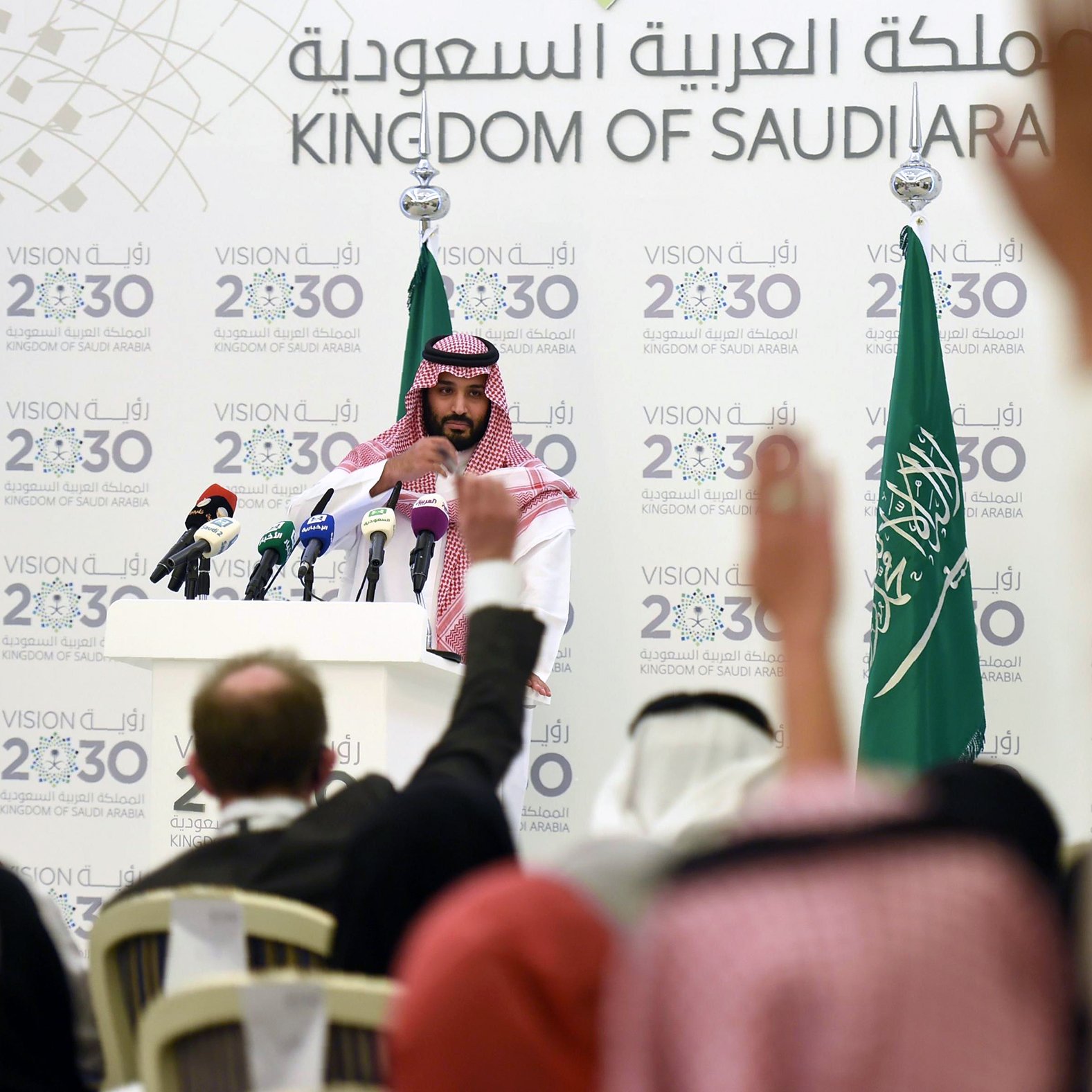 L'image montre une conférence de presse organisée en Arabie Saoudite. Un homme, vêtu d'une robe traditionnelle saoudienne et d'un headdress, se tient devant un podium avec plusieurs microphones. En arrière-plan, on peut voir des drapeaux saoudiens ainsi qu'un panneau affichant des informations sur la Vision 2030 de l'Arabie Saoudite. Plusieurs personnes sont présentes, levant les mains, probablement pour poser des questions. L'atmosphère semble sérieuse, centrée sur des discussions importantes liées à l'avenir du pays.