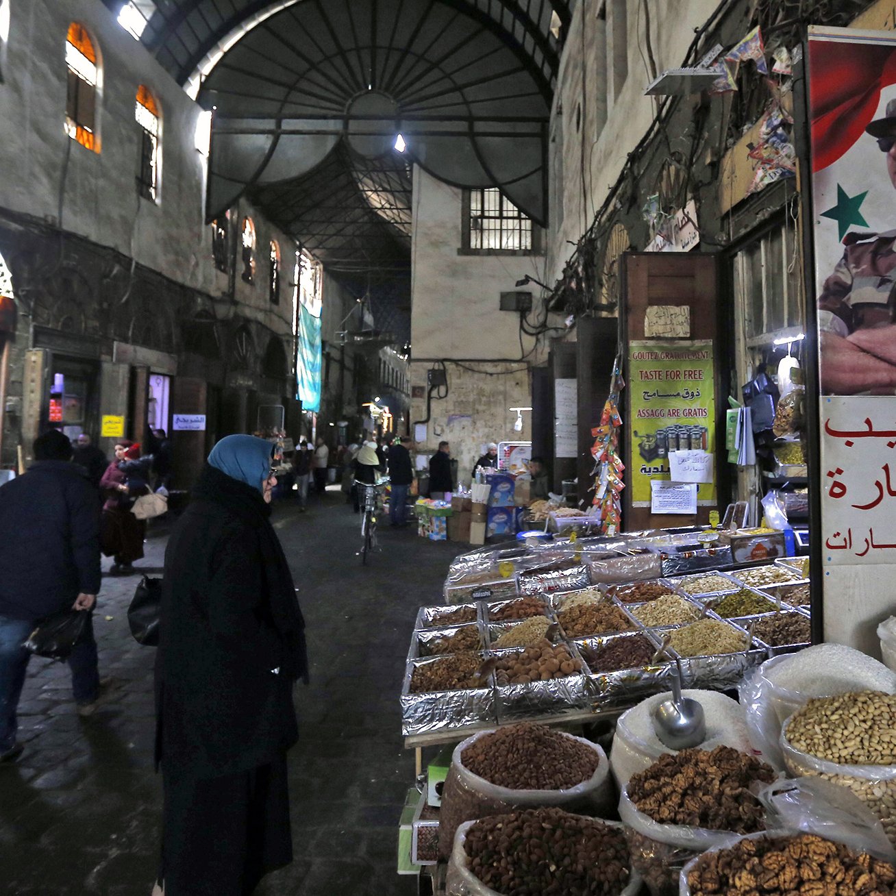 L'image montre une scène d'un souk ou d'un marché typique, probablement en Syrie. On aperçoit des étals avec des épices, des fruits secs, et d'autres produits en vente. Les murs de la rue sont en pierre et semblent anciens, créant une ambiance historique. Des personnes, dont certaines portent des vêtements traditionnels, se déplacent dans la rue, tandis qu'une affiche avec un symbole militaire est accrochée à un mur, ajoutant un élément de contexte culturel et politique à la scène. La lumière faible et les ombres accentuent l'ambiance conviviale et animée du marché.