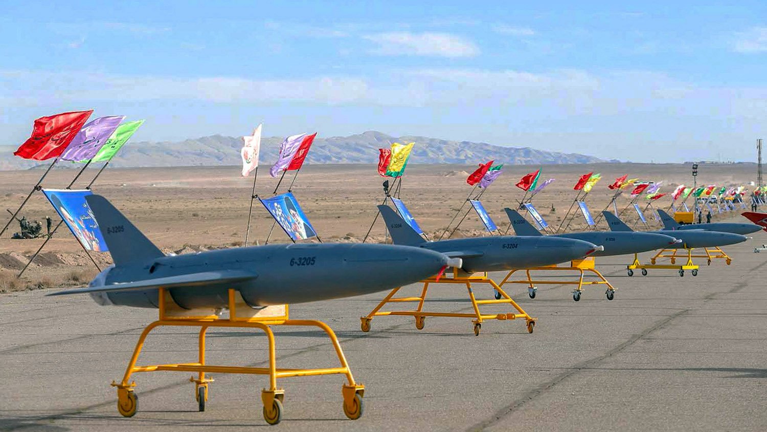 تظهر الصورة مجموعة من الطائرات المسيرة (الدرون) متراصة على مدرج، حيث كل طائرة تحمل علمًا مختلفًا ملونًا. الخلفية تضم مناظر طبيعية صحراوية مع جبال في الأفق، مما يدل على بيئة جافة. التصميم العام للطائرات يبدو حديثًا، مما يعكس تطور التكنولوجيا العسكرية.