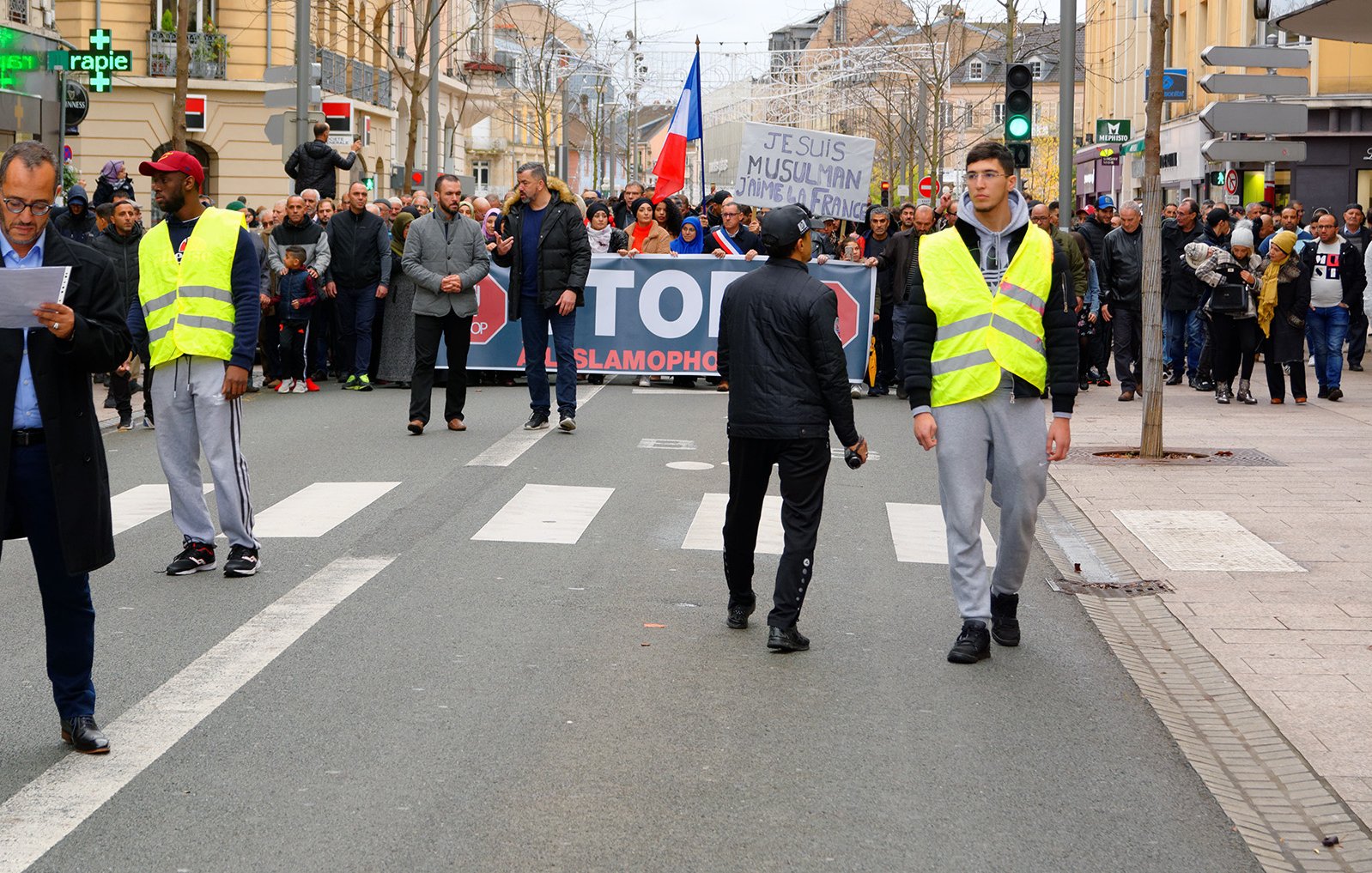 الصورة تظهر مجموعة من الناس يتظاهرون في شارع. في المقدمة، هناك شخصان يرتديان سترة عاكسة، ويبدو أنهما يجريان أو يقومان بمراقبة المظاهرة. في الخلفية، يمكن رؤية لافتة كبيرة مكتوب عليها "STOP ISLAMOPHOBIE"، مما يشير إلى أن المظاهرة تتعلق بمناهضة الإسلاموفوبيا. المتظاهرون يحملون لافتات ويبدو أنهم يتجمعون بشكل منظم. الجو العام يعكس النشاط والحيوية.