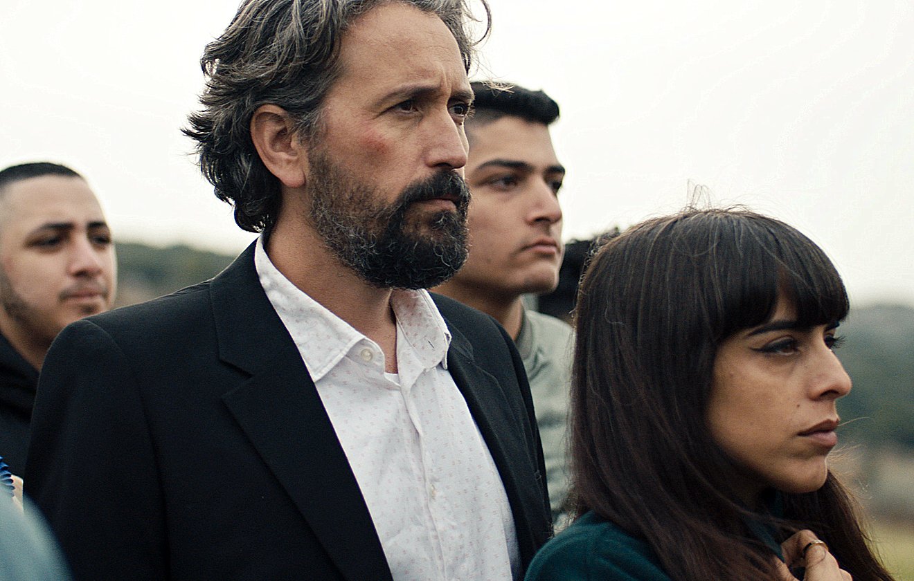 L'image montre un groupe de personnes rassemblées, avec un homme au premier plan, portant un costume noir et une chemise blanche. Il a les cheveux gris et une barbe. À côté de lui se trouve une femme avec des cheveux longs et bruns, qui semble pensive. En arrière-plan, d'autres personnes sont présentes, affichant des expressions sérieuses. L'ambiance général semble solennelle.