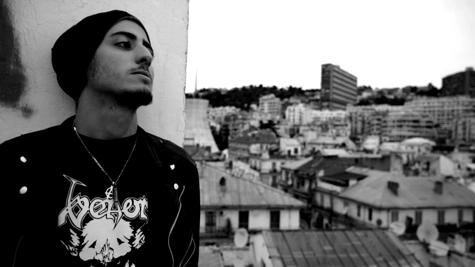 L'image montre un jeune homme sur un toit, regardant au loin. Il porte un t-shirt noir avec un motif graphique et une veste. Son look est accentué par un bonnet. En arrière-plan, on peut voir une vue de la ville avec des bâtiments et des toits, le tout dans des teintes en noir et blanc qui donne une atmosphère mélancolique. La scène évoque une ambiance urbaine et contemplative.