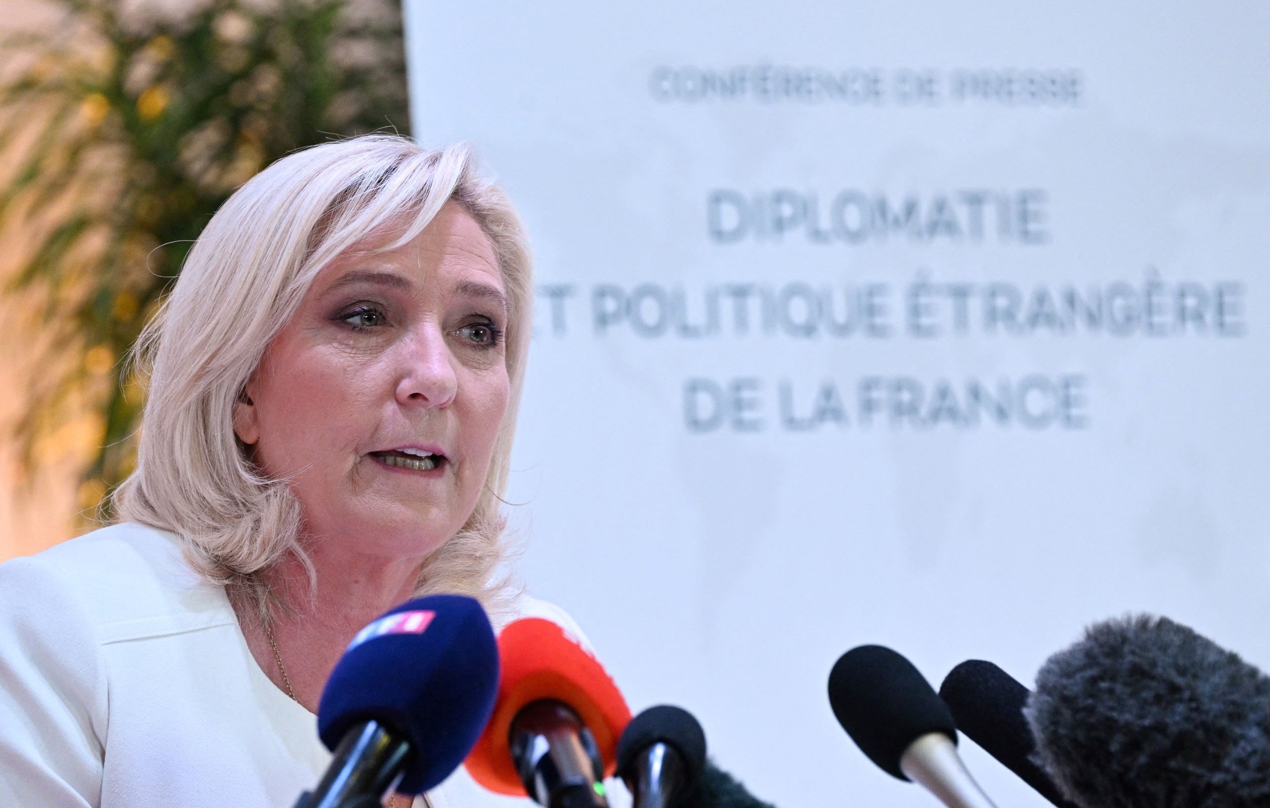 L'image montre une femme qui s'exprime lors d'une conférence de presse. Elle a des cheveux blonds et porte une veste claire. Au fond, on peut voir un panneau qui indique "CONFERENCE DE PRESSE" ainsi que le titre "DIPLOMATIE ET POLITIQUE ÉTRANGÈRE DE LA FRANCE". Plusieurs microphones sont disposés devant elle, suggérant qu'elle s'adresse aux médias. L'ambiance semble formelle et professionnelle.