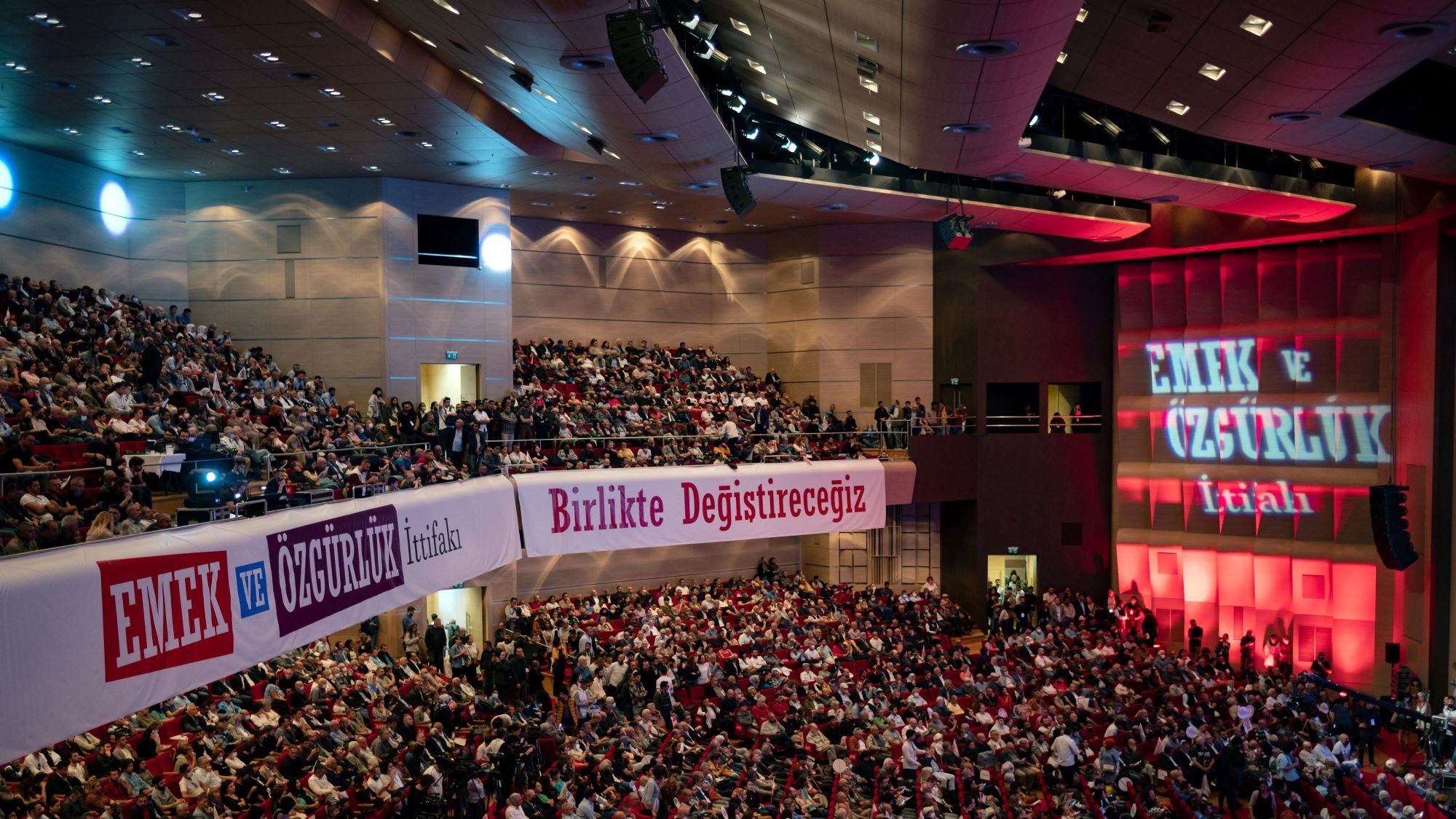 در این تصویر، صحنه‌ی یک سالن بزرگ کنفرانس با تعدادی بسیار زیاد از افراد در حال نشستن و تماشا دیده می‌شود. سالن به خوبی روشن است و روی دیوارهای آن نوشته‌هایی به زبان ترکی وجود دارد. یکی از این نوشته‌ها به صورت برجسته "EMEK VE ÖZGÜRLÜK" (به معنی "کار و آزادی") است. فضا به نظر می‌رسد مربوط به یک همایش یا تجمع سیاسی باشد که در آن مردم به موضوعاتی مهم توجه دارند. در پس‌زمینه، نورپردازی خاصی وجود دارد که بر احساسات و فضای کنفرانس تأکید می‌کند.