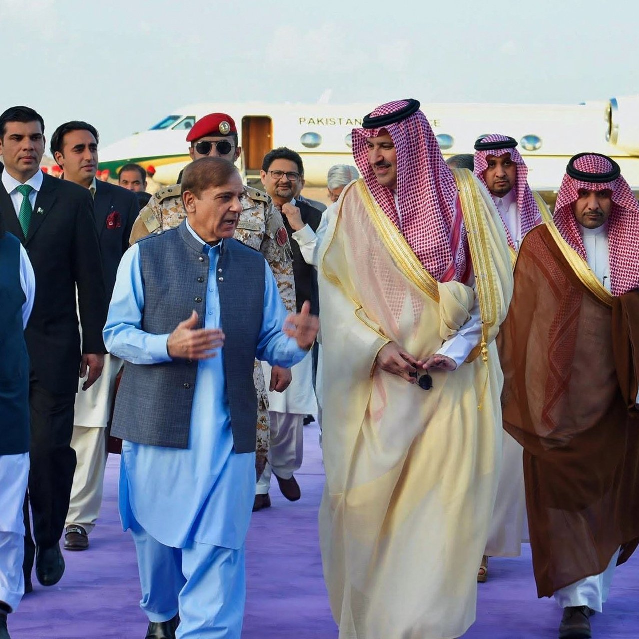 The image depicts a group of men walking together, likely during a formal event or diplomatic visit. They are dressed in traditional attire, with some wearing long robes and others in suits. The setting appears to be an airport or other official venue, indicated by the presence of a private jet in the background. The men seem engaged in conversation, and the atmosphere suggests camaraderie or collaboration. The purple carpet hints at a ceremonial welcome.