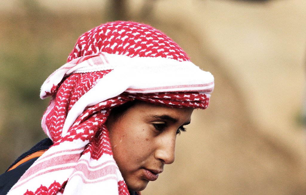 در این تصویر، یک فرد با چهره‌ای متعلق به سن جوانی دیده می‌شود. او یک کلاهی با طرح قرمز و سفید بر سر دارد که به شیوه‌ای سنتی پوشیده شده است. چهره‌اش به سمت پایین متمایل است و نگاهی تفکرآمیز دارد. پس‌زمینه تصویر مبهم است و توجه بیشتر بر روی شخص متمرکز شده است. این تصویر حس آرامش و تمرکز را منتقل می‌کند.