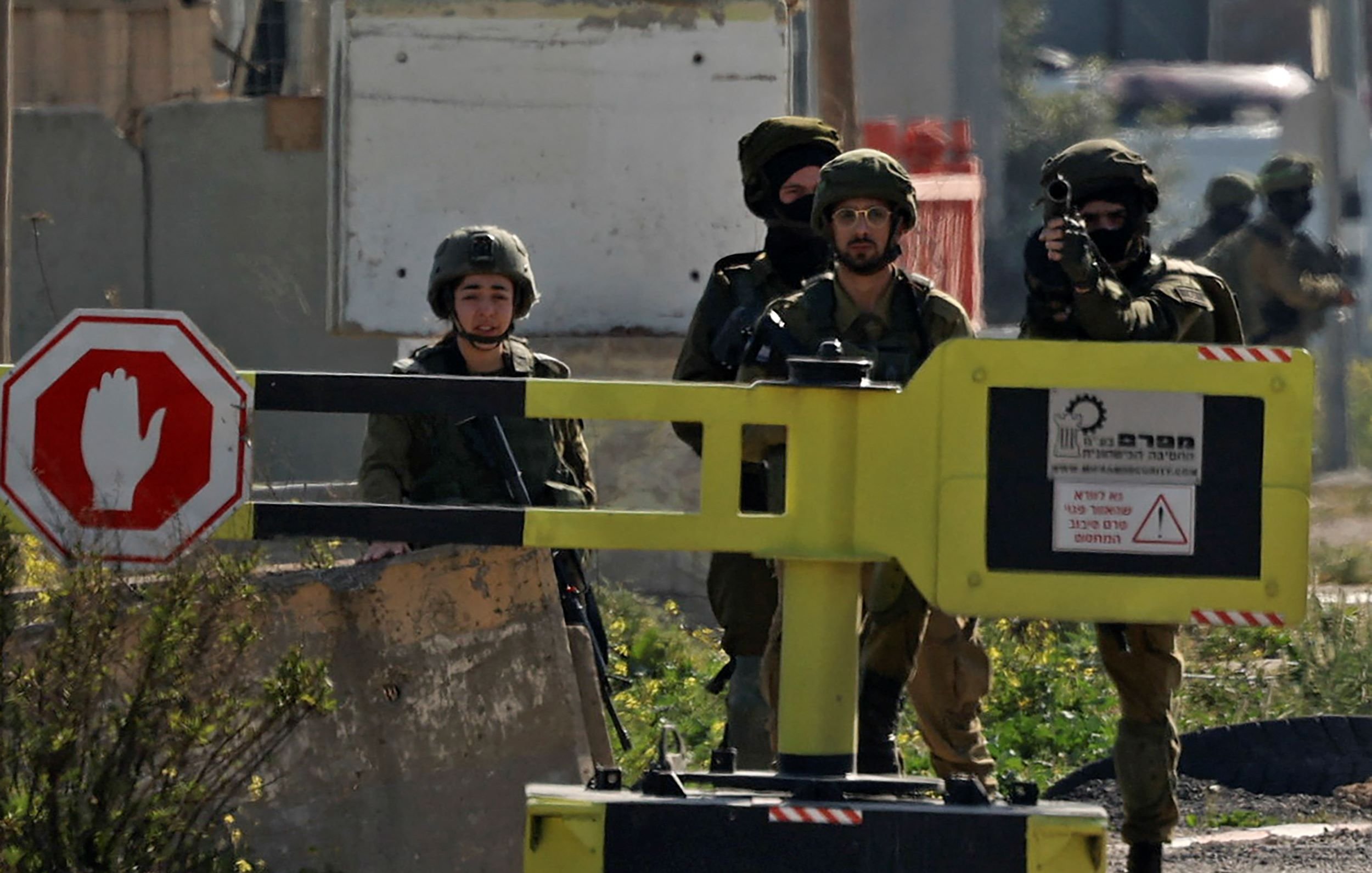 L'immagine mostra un gruppo di soldati in un ambiente urbano. Sono vestiti con uniformi militari e indossano elmetti. Due soldati sono in piedi vicino a un blocco stradale giallo con un segnale di fermo e il gesto di "stop". Nel gruppo, uno dei soldati sta puntando un'arma in direzione non visibile. Il contesto sembra suggerire un'operazione di sicurezza o un controllo di accesso. Sullo sfondo, ci sono strutture edilizie e segni di una zona di conflitto.