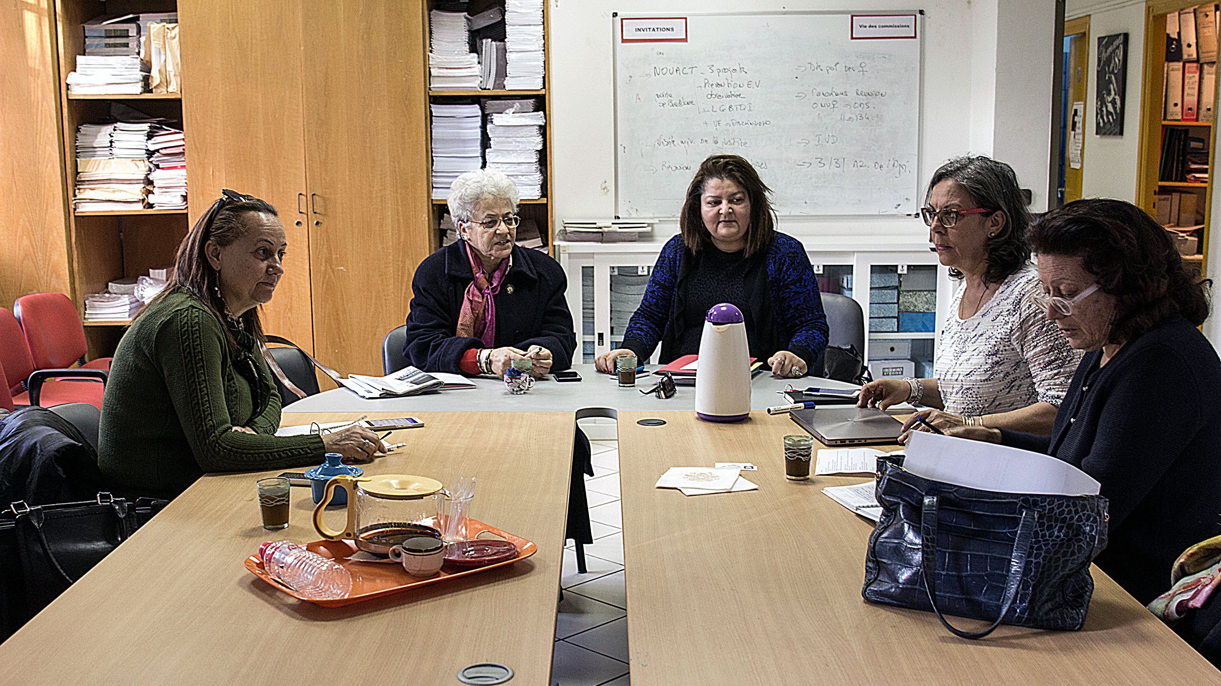 L'image montre un groupe de femmes assises autour d'une table dans une pièce qui semble être un espace de réunion. Elles sont engagées dans une discussion ou une activité collaborative. Sur la table, on peut voir des carnets, des verres et un thermos. L'environnement est simple, avec des étagères remplies de dossiers et de documents en arrière-plan. Les femmes semblent concentrées et impliquées dans leur échange.