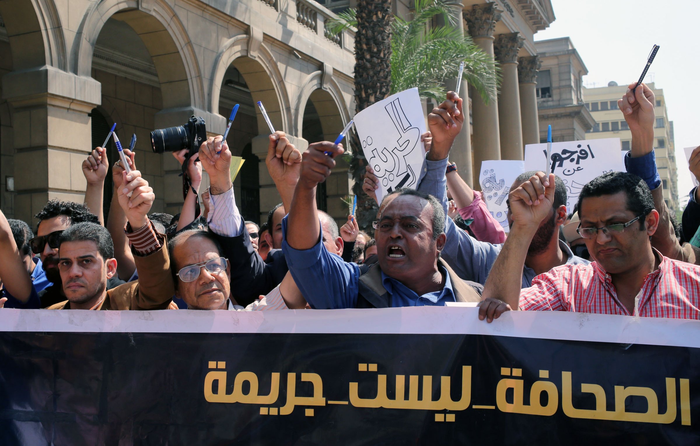 L'image montre une manifestation avec un groupe de personnes levant les bras et tenant des pancartes. Ils semblent exprimer leur mécontentement ou revendiquer des droits, notamment en rapport avec la liberté de la presse. L'atmosphère est énergique et déterminée, avec des visages montrant des émotions de passion et de revendication. À l'arrière-plan, on peut voir des bâtiments qui indiquent un environnement urbain. Le slogan sur la banderole semble défendre une position sur la presse, suggérant que celle-ci ne devrait pas être considérée comme un crime.