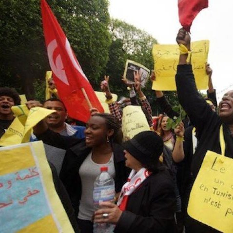 L'image montre une manifestation dans une rue animée, avec des gens qui tiennent des pancartes et des drapeaux. Une personne au centre, habillée en noir, lève un panneau sur lequel est écrit "Le Racisme est un délit et un crime en Tunisie". Les manifestants semblent exprimer des préoccupations concernant le racisme. L'ambiance est vibrant et engagée, avec des participants qui chantent ou crient des slogans. Les éléments visuels suggèrent une mobilisation sociale pour des droits et la justice.