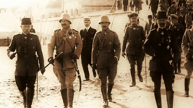 The image depicts a historical scene with a group of military personnel walking together in a line. They are dressed in formal military uniforms and accessories, including hats and medals, indicating a high rank or importance. The setting appears to be outdoors, possibly on a dirt or gravel path, with tents in the background suggesting a military camp. The overall tone of the photograph has a sepia or monochrome effect, conveying a sense of history. The individuals appear to be engaged in conversation or in a social setting, reflecting camaraderie among them.
