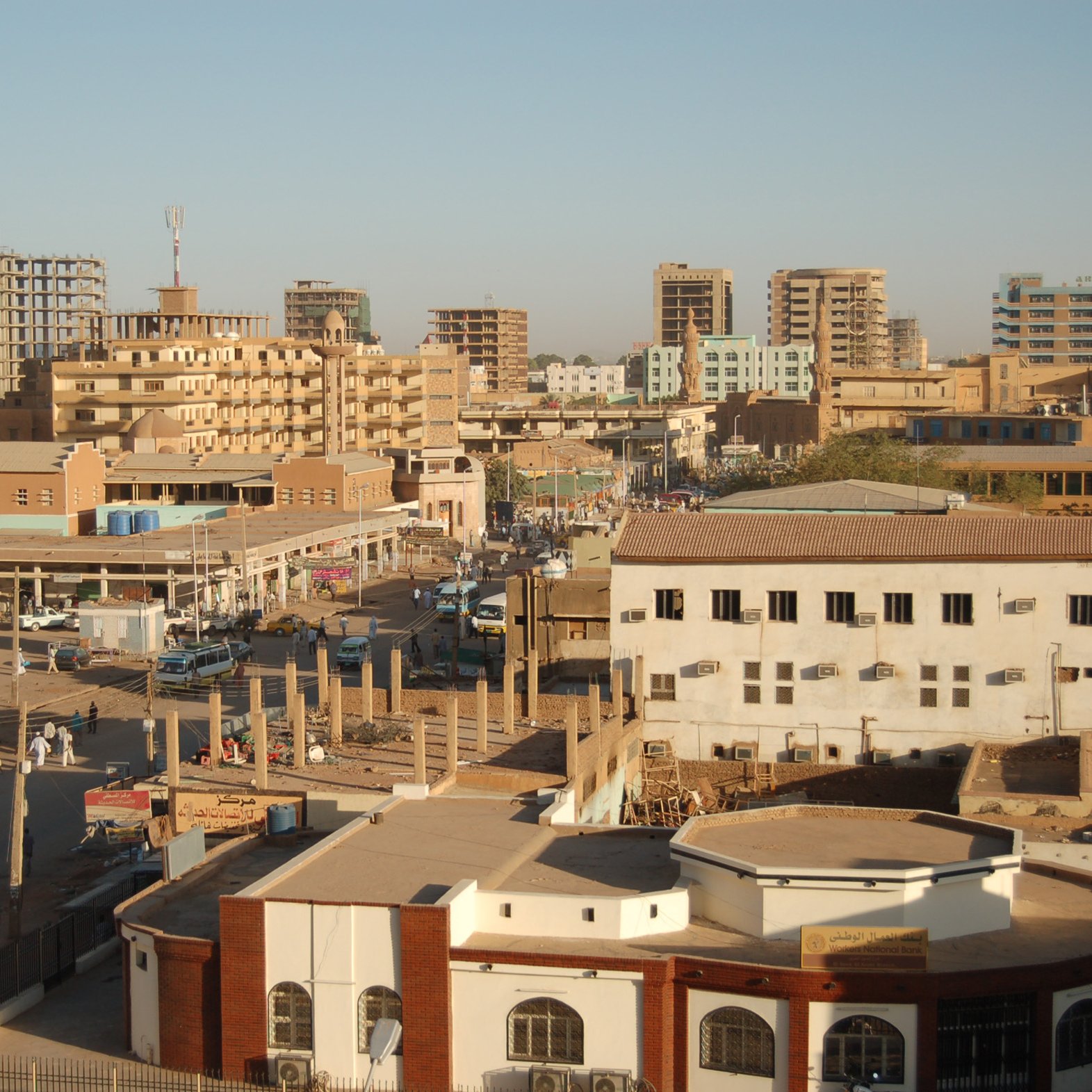 الصورة تظهِر مشهداً حضرياً لمدينة تحتوي على مبانٍ متعددة الطوابق، وبعض المباني قيد الإنشاء. تُظهر الشوارع المزدحمة بالسيارات والمارة، بالإضافة إلى وجود أسواق محلية وأعمال تجارية. السماء صافية، وهناك ضوء شمس ساطع، مما يوحي بأن الوقت نهار. الألوان في الصورة تتراوح بين درجات البني والرمادي، مما يعكس طابع المدينة.