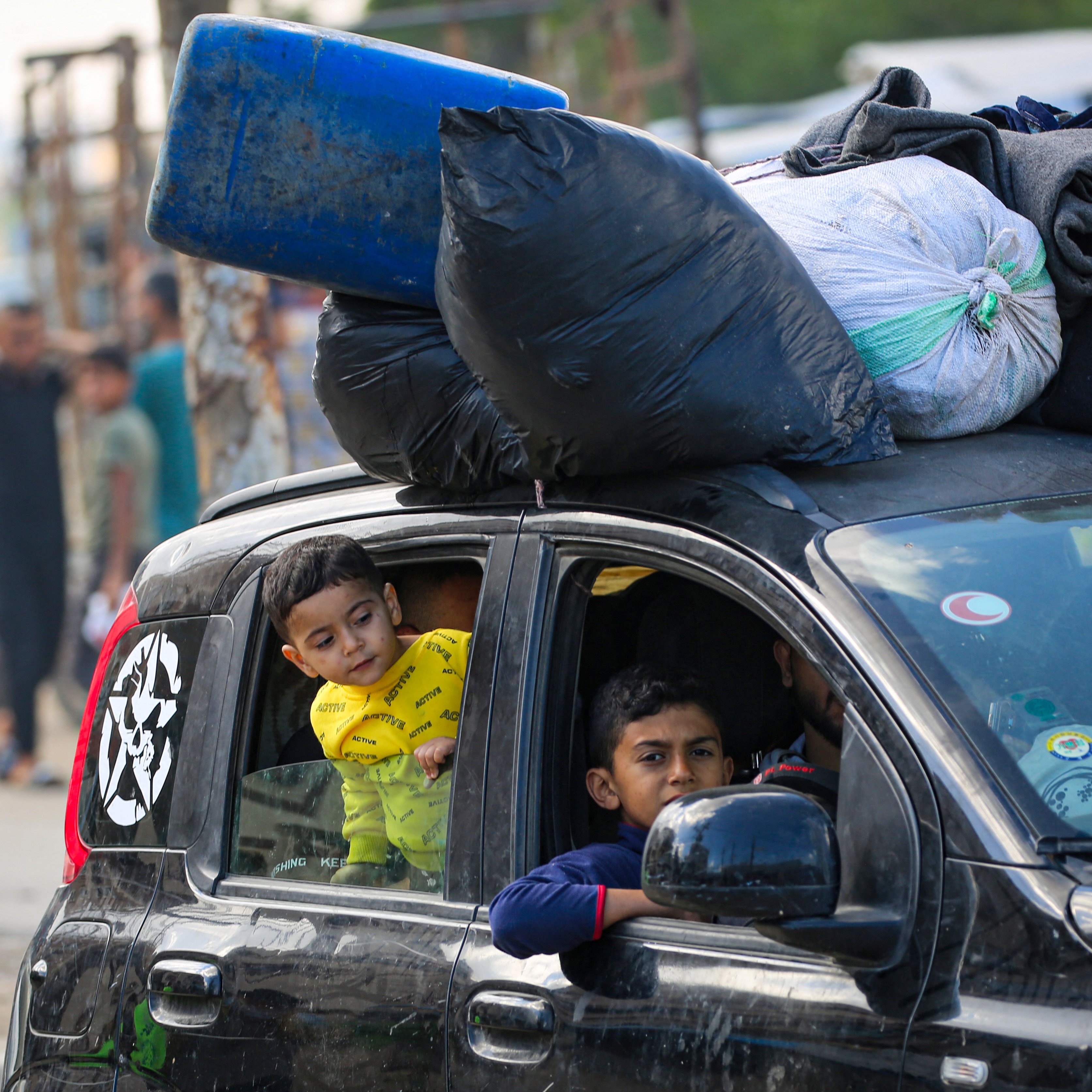 Sur cette image, on voit une voiture noire avec plusieurs objets attachés sur le toit, notamment des sacs et un bidon bleu. À l'intérieur du véhicule, il y a deux enfants. L'un des enfants, portant un vêtement jaune avec un imprimé, regarde par la fenêtre de la voiture. L'autre enfant, assis à l'avant, regarde également dehors. En arrière-plan, il y a plusieurs personnes qui semblent se déplacer, ainsi qu'un environnement urbain sans détails précis.