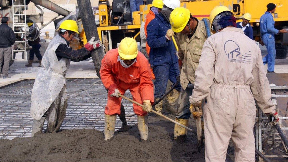 L'image montre plusieurs ouvriers en train de travailler sur un chantier de construction. Ils portent des casques de sécurité jaunes et des vêtements de protection, tels que des combinaisons et des bottes en caoutchouc. L'un des travailleurs utilise une pelle pour manipuler du béton, tandis que d'autres semblent s'occuper de l'installation ou de la préparation du béton. En arrière-plan, on peut voir des machines de chantier et des équipements. L'atmosphère est active, indiquant un environnement de travail intense.