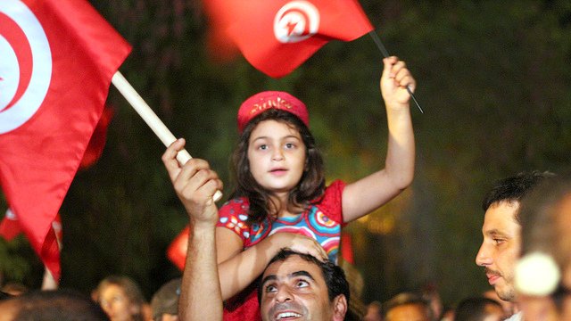 L'image montre une scène de célébration en Tunisie. Une jeune fille, vêtue d'une robe colorée, est assise sur les épaules d'un homme qui sourit. Ils brandissent des drapeaux tunisiens. En arrière-plan, on aperçoit une foule joyeuse, créant une atmosphère festive. Les expressions des visages reflètent la joie et l'engagement.