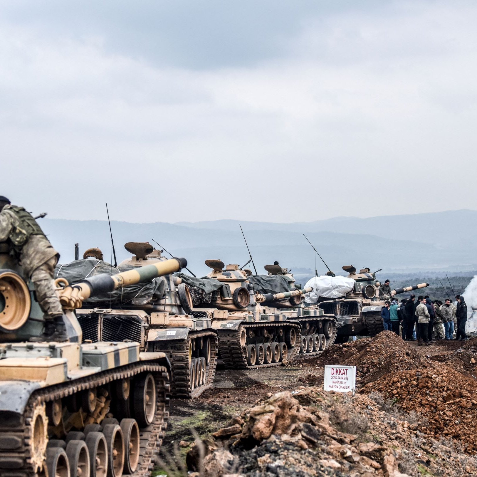 L'image montre une ligne de chars militaires, probablement en position de déploiement ou d'entraînement. Les chars sont recouverts de bâches et semblent être stationnés sur un terrain accidenté, entourés de collines. Au loin, on peut apercevoir des militaires rassemblés autour de ce qui semble être un poste de commandement ou d'observation. Il y a également des signes ou des marquages au sol, et l'atmosphère paraît chargée, avec une légère brume ou fumée dans l'air.
